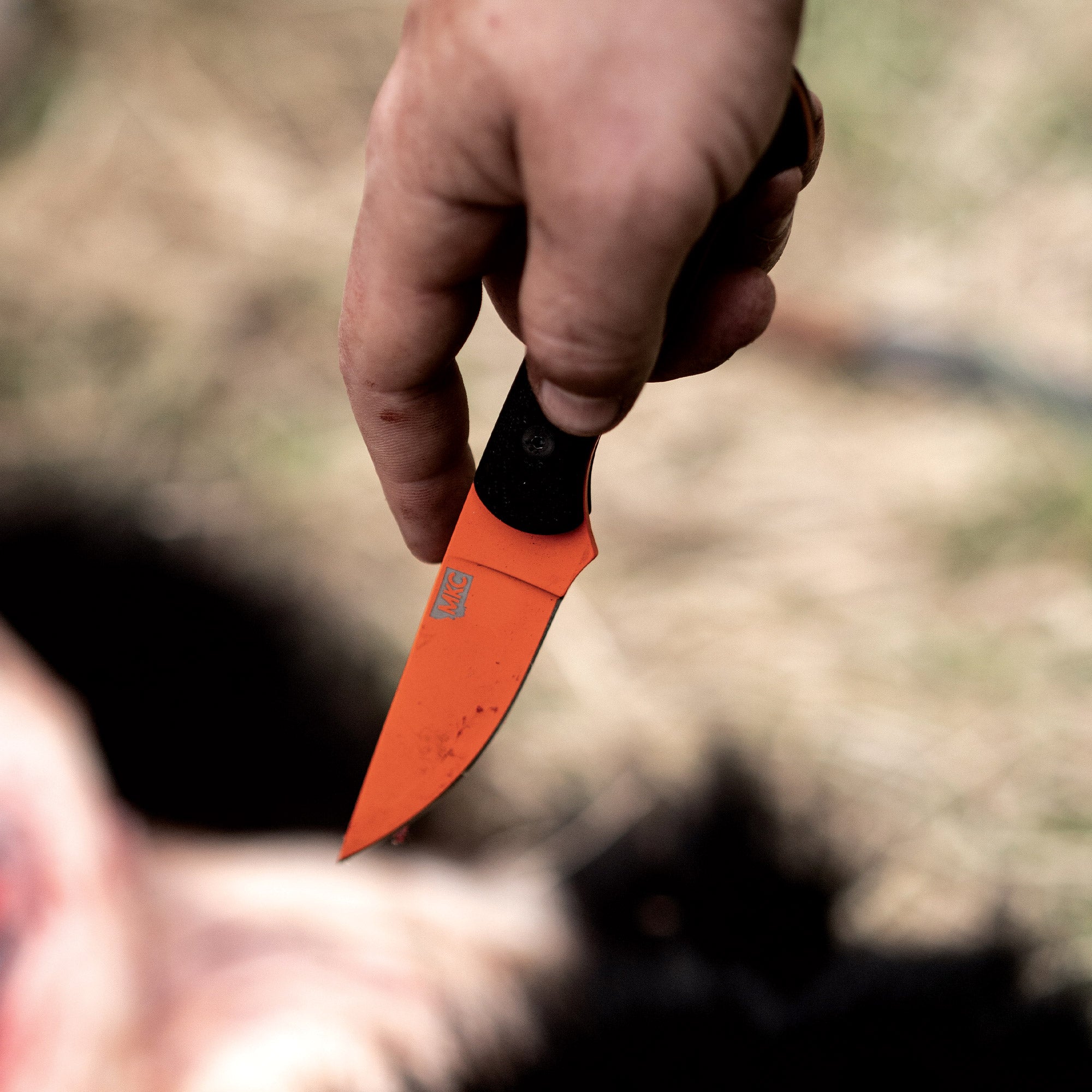 Alt text: A close-up view of a hand holding a BLACKFOOT 2.0 fixed blade knife with a blaze orange Cerakote™ coating and black grip, displayed outdoors.