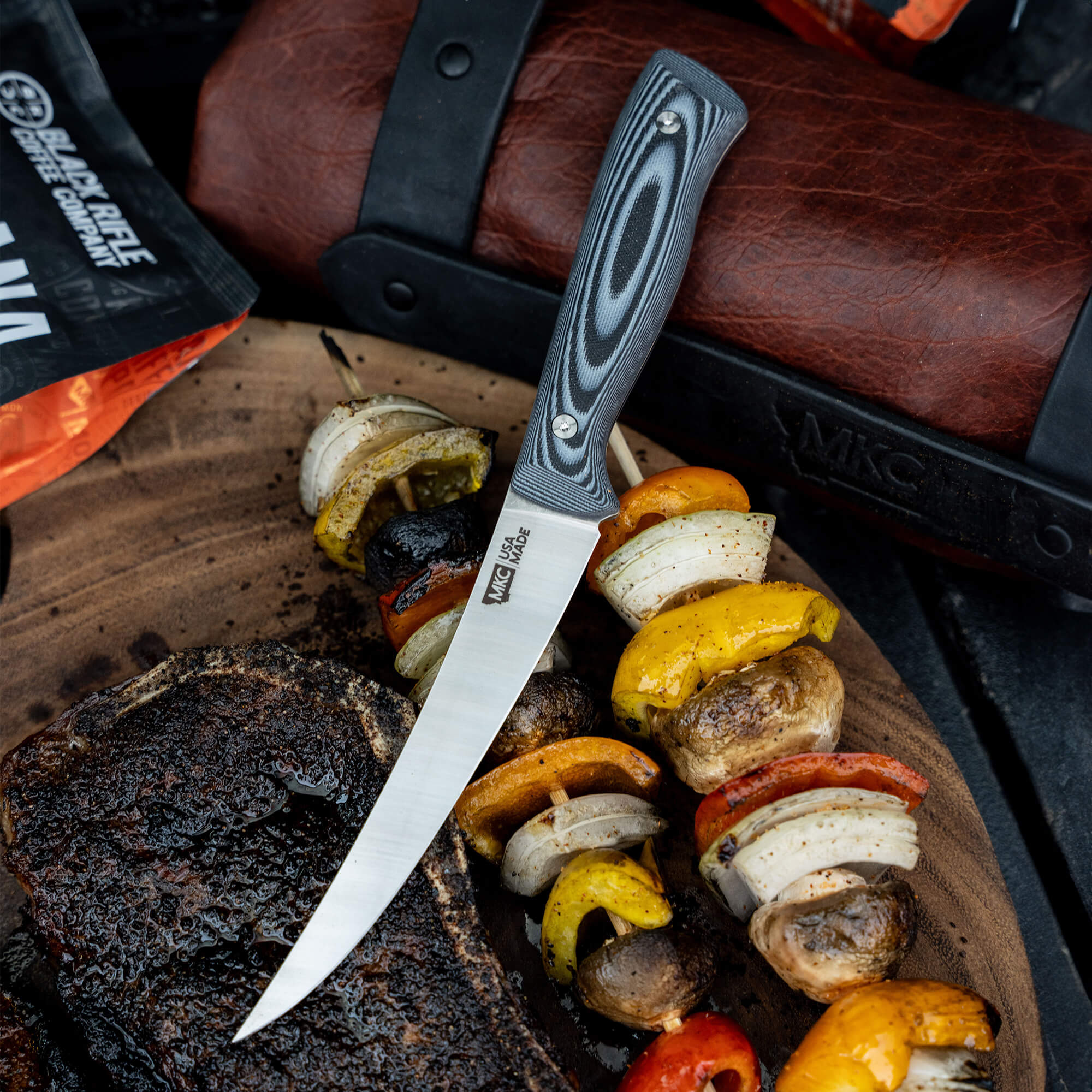 Alt-text: "Bearded Butchers boning butcher knife with a intricately designed gray and black handle, displayed on a wooden board, piercing through a freshly grilled assortment of vegetables and steak, with a leather case in the background, highlighting its sharp
