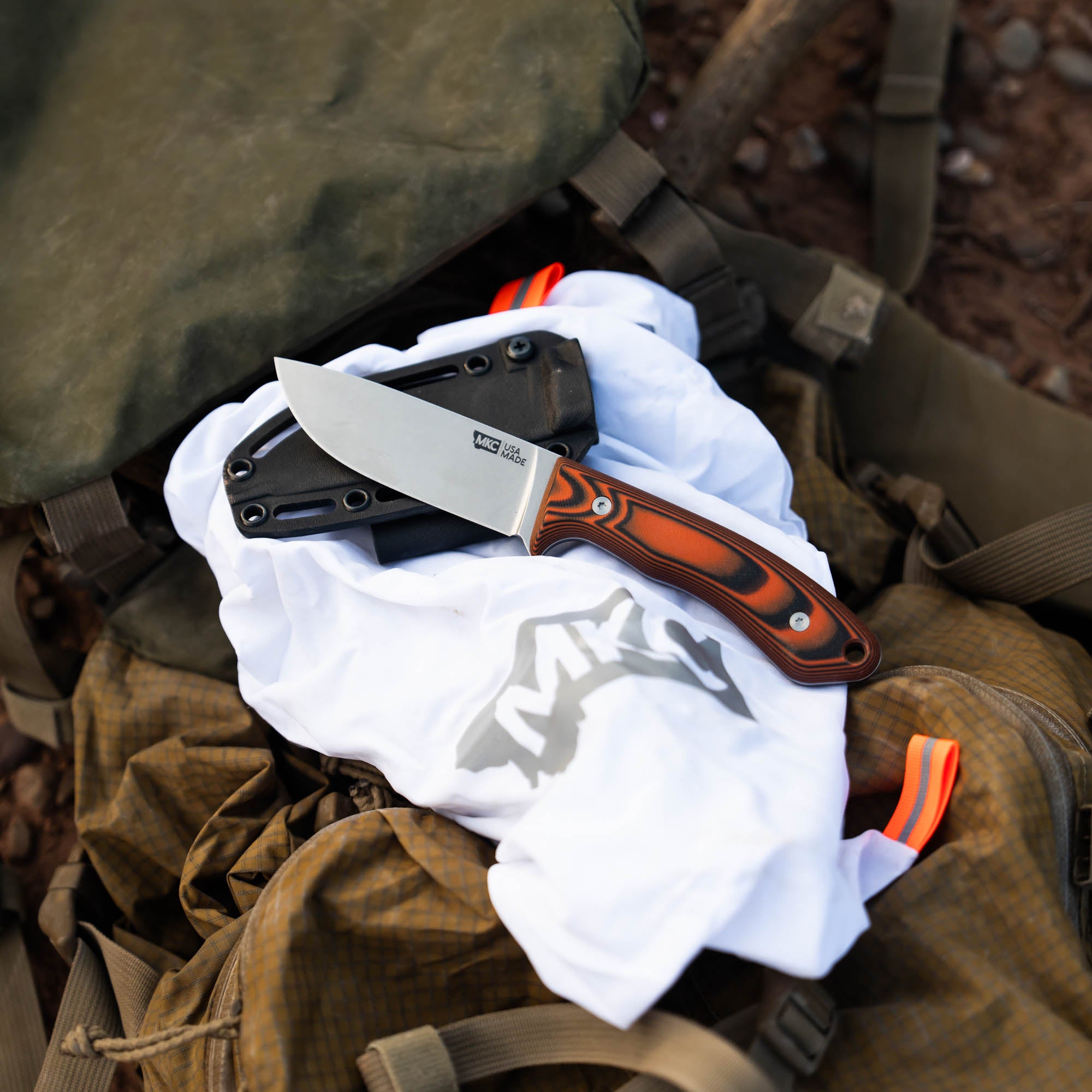 MKC Mule Deer Knife in orange and black, resting on a white shirt inside a backpack, showcasing its durable design.