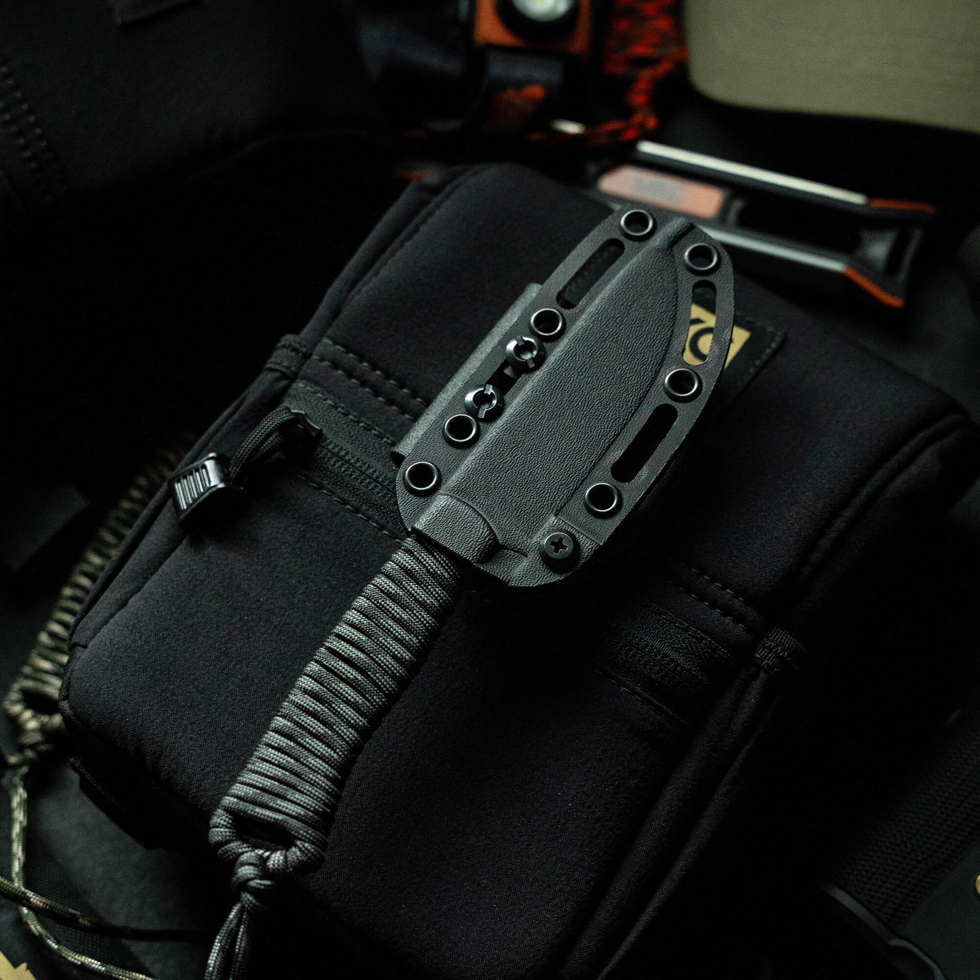 Alt text: "Wild Sheep Foundation Stoned Goat 2.0 knife, featuring a textured black handle and metal sheath, securely attached to a tactical black bag, with rugged outdoor equipment in the background, indicating the knife's professional and robust design