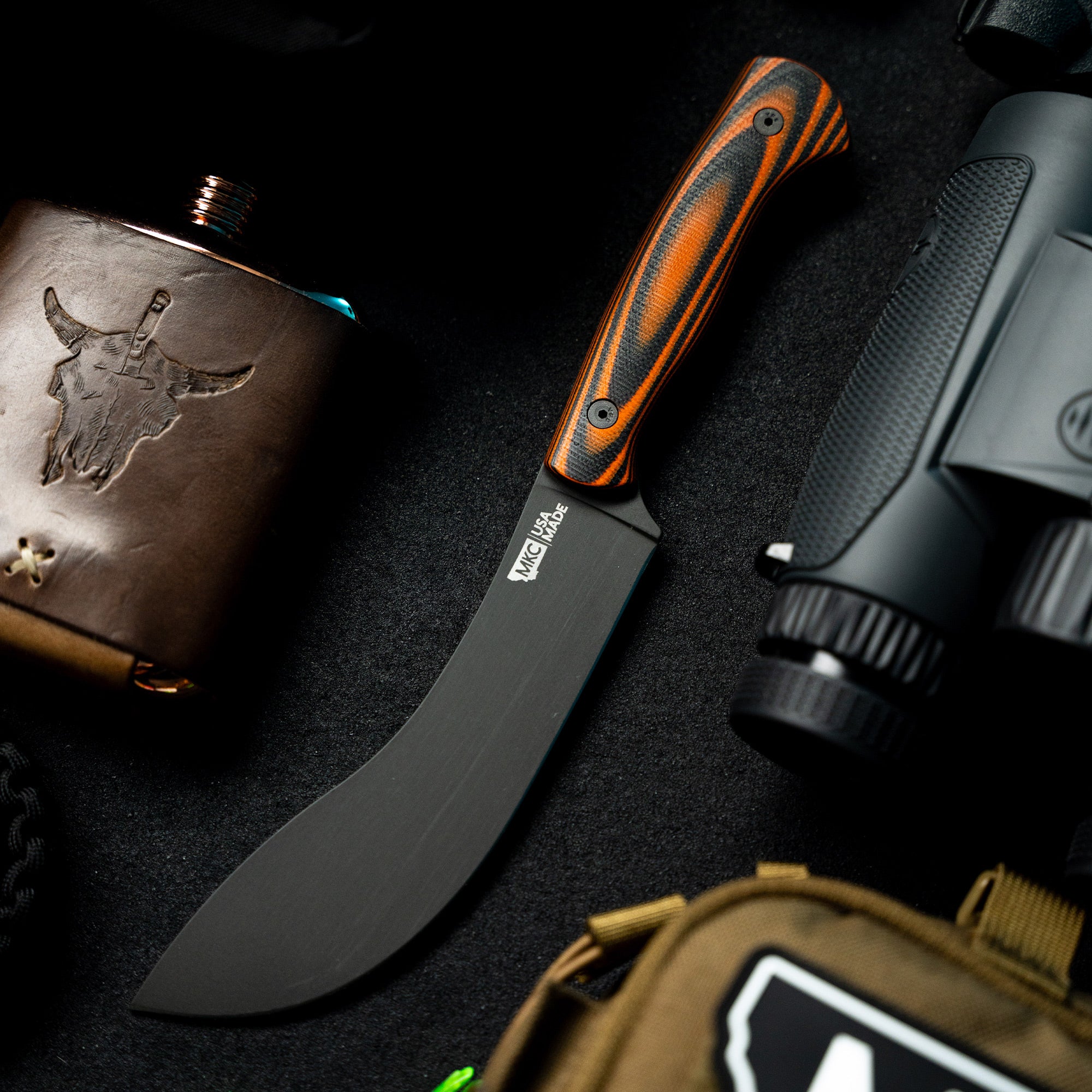 Alt text: High-quality image of a BEARTOOTH Pro Skinning Blade with an orange and black handle, prominently displayed alongside outdoor gear including a leather flask and a tactical binocular, all set on a dark textured background.