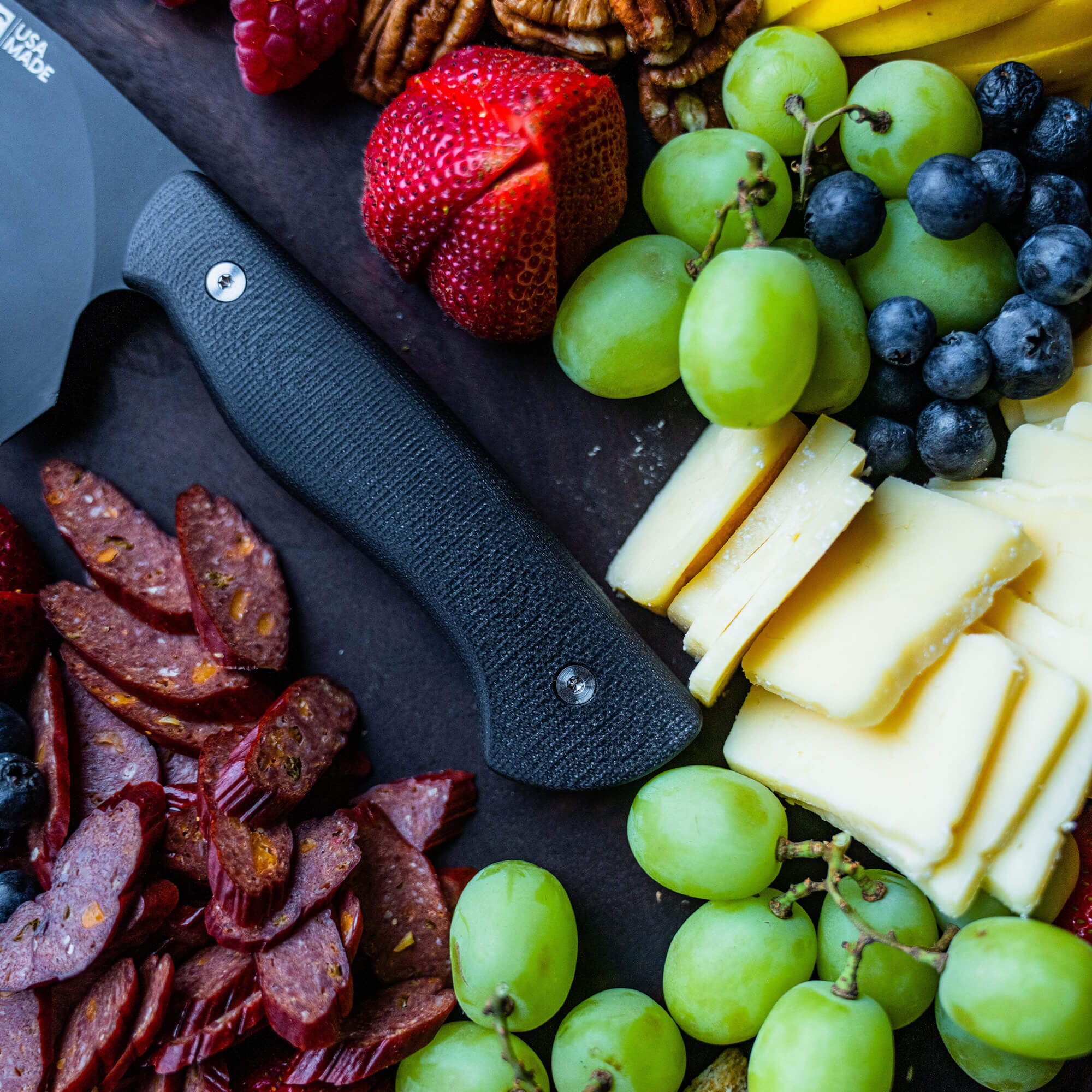 Black-handled Bighorn Chef knife on a charcuterie board with assorted meats, cheeses, and fruits including strawberries, grapes, and blueberries.