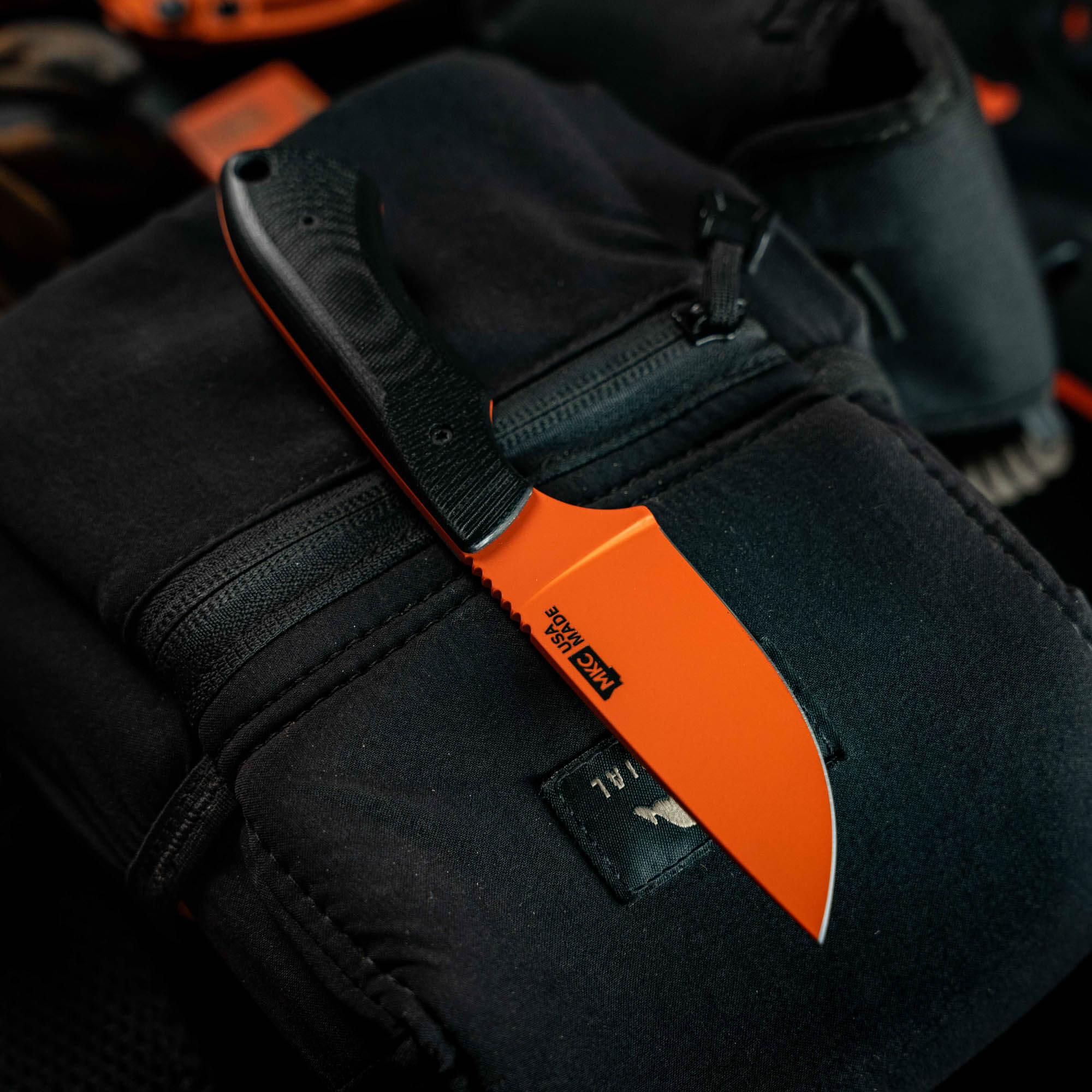 MKC Elk Knife with Blaze Orange Cerakote finish, featuring a black textured handle, resting on a dark fabric background.