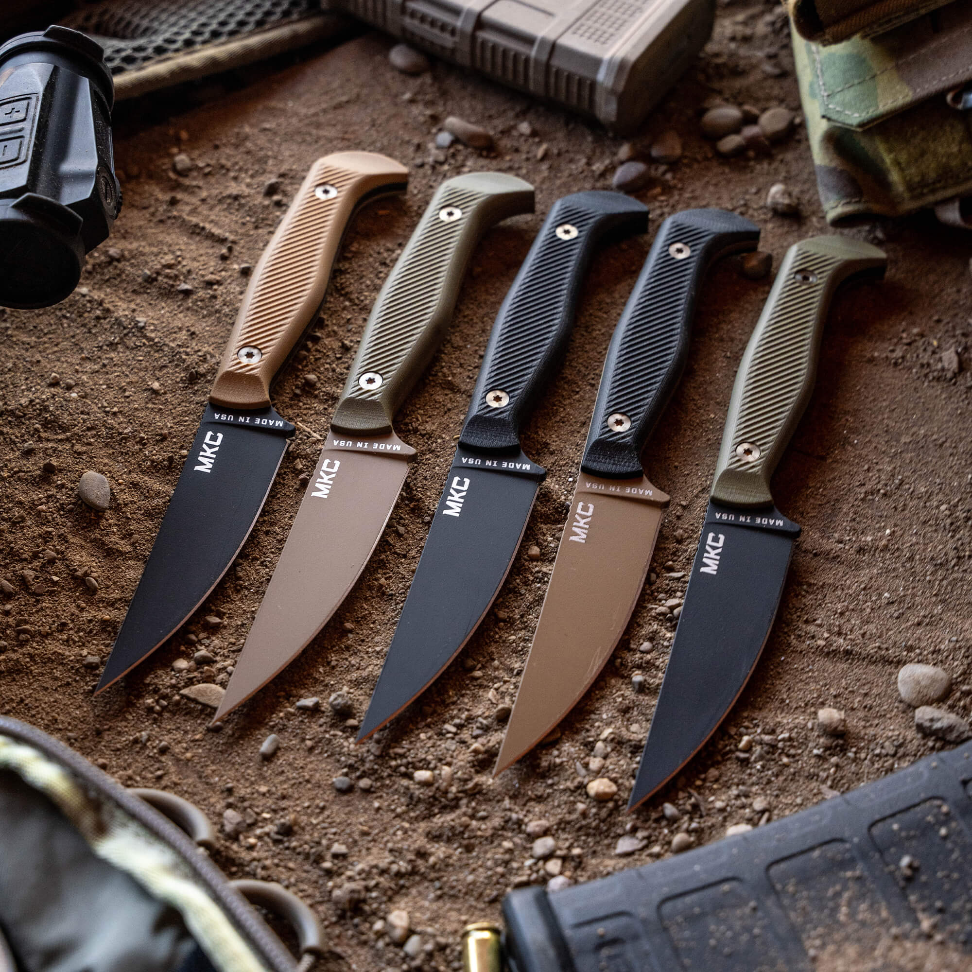 Alt text: "Array of five MKC Battle Goat fixed-blade knives displayed on a sandy surface, featuring various handle colors including black and tan, with the MKC logo prominently displayed on each blade."