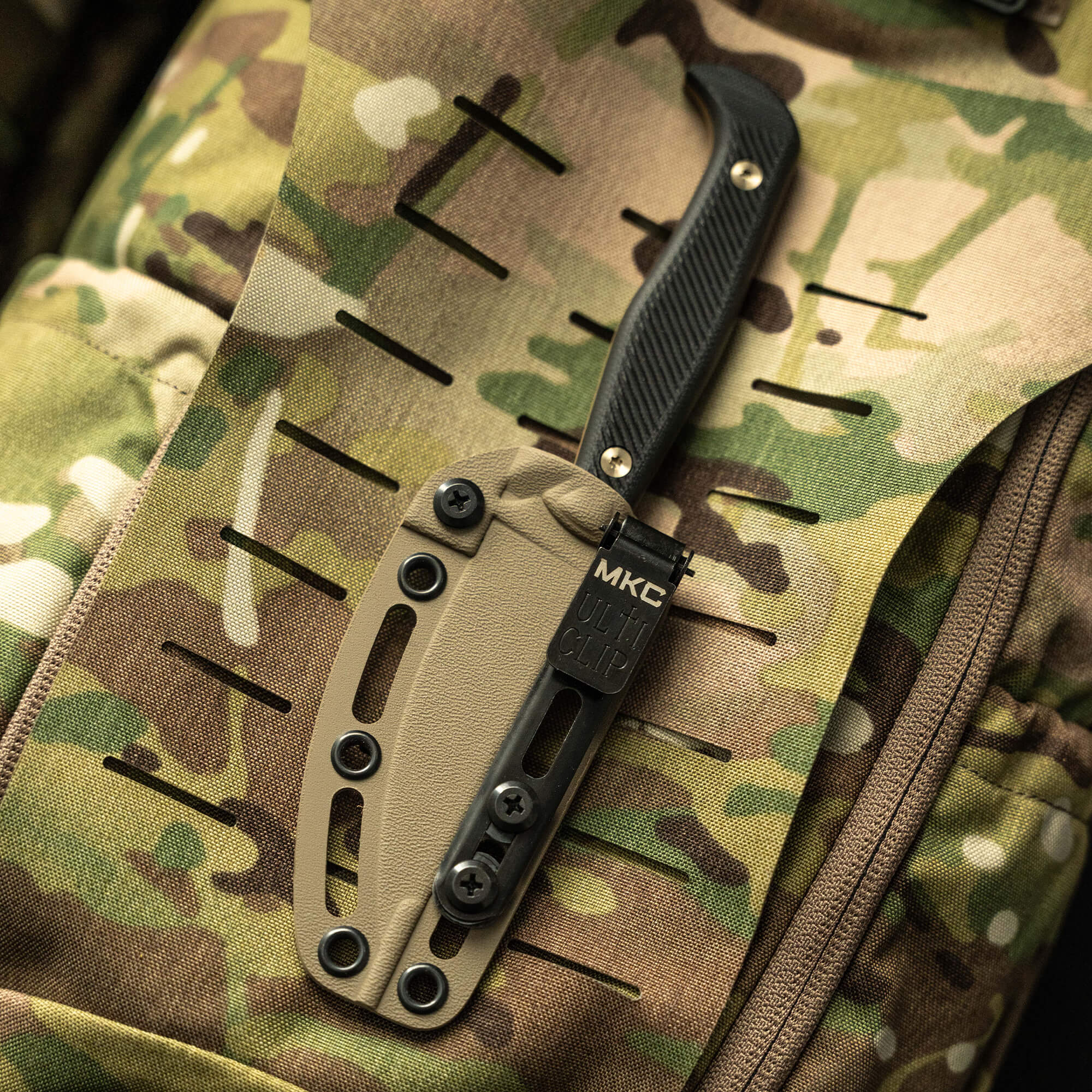 Alt text: A coyote black Battle Goat tactical knife attached to a camouflage military backpack, featuring a sturdy black handle, tan sheath with black metal clip inscribed with "MKC ULT CLIP".