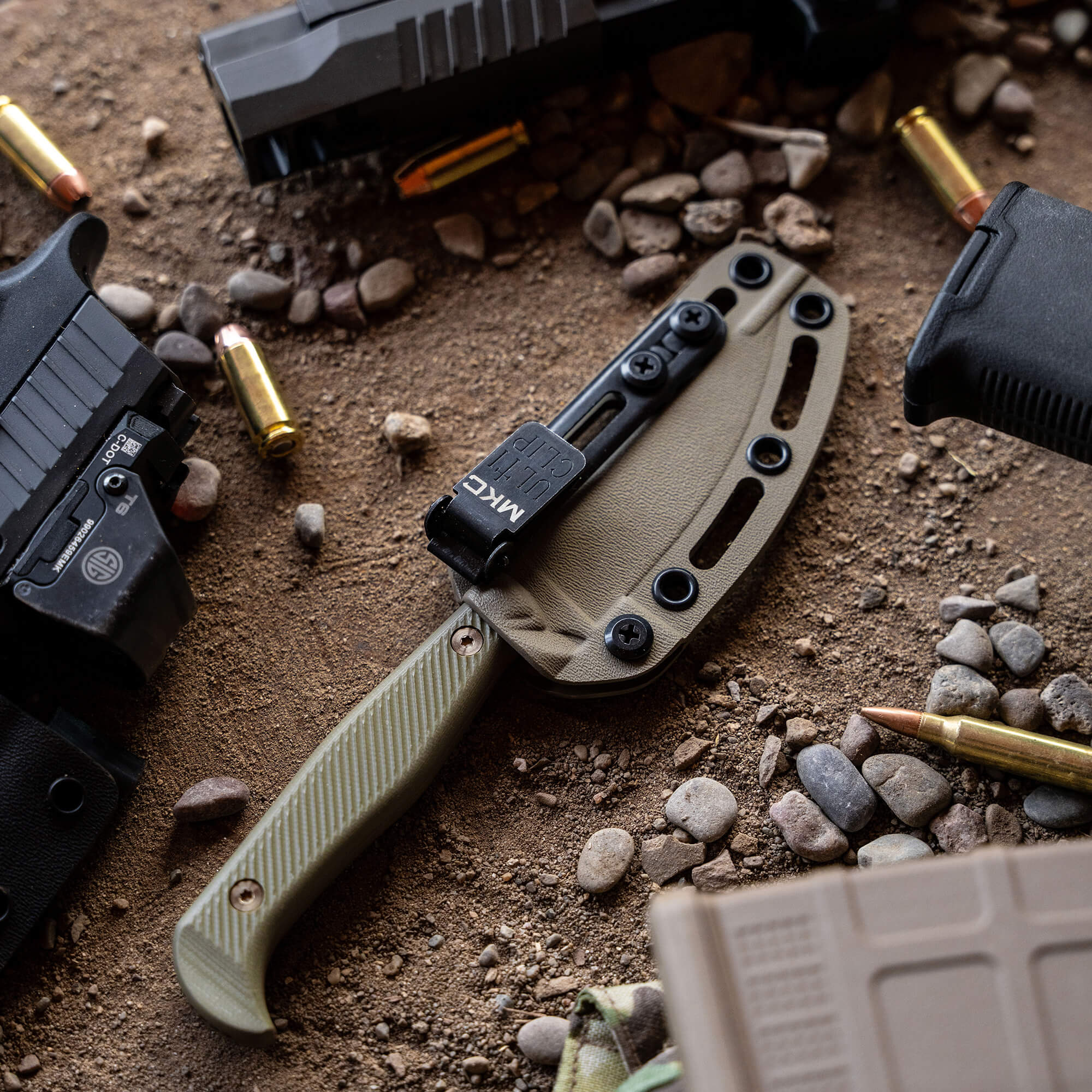 Alt text: "Tactical knife 'Battle Goat' in coyote olive drab color with a tan sheath, positioned on a sandy ground surrounded by scattered ammunition and parts of tactical gear."