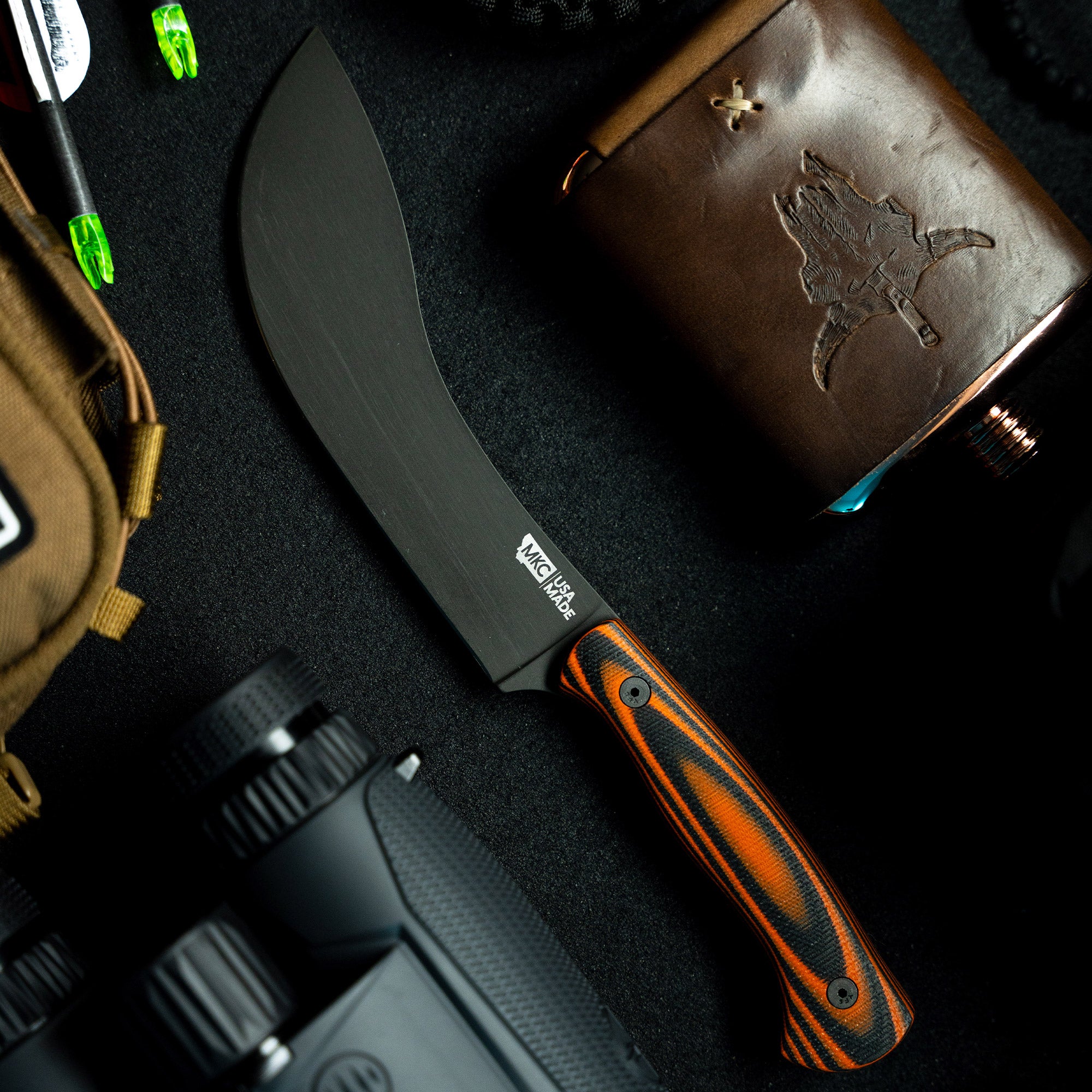 Alt text: "BEARTOOTH Pro Skinning Blade with an orange and black striped handle prominently displayed on a dark textured surface, accompanied by outdoor gear including a camera, flask with embossed map design, and a tactical backpack."