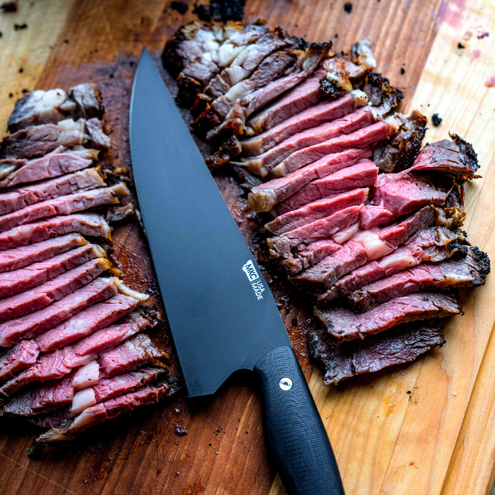 Alt text: "A large, black Bighorn Chef knife beside perfectly sliced, medium-rare brisket on a wooden cutting board, showcasing the knife's sharpness and professional quality in a culinary setting."