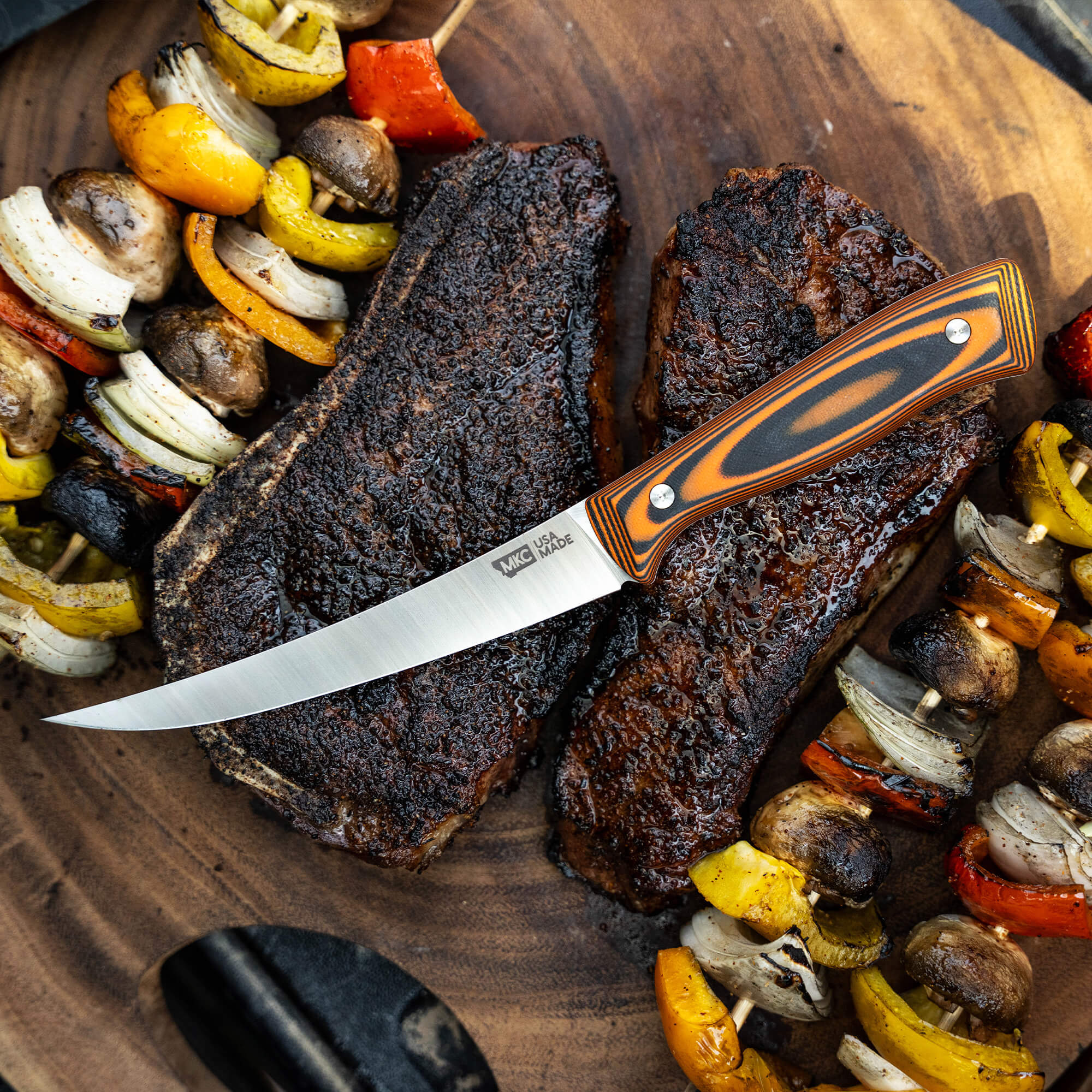 Alt text: "Bearded Butchers boning butcher knife with an orange and black handle, displayed on a wooden board next to cooked brisket and grilled vegetables."