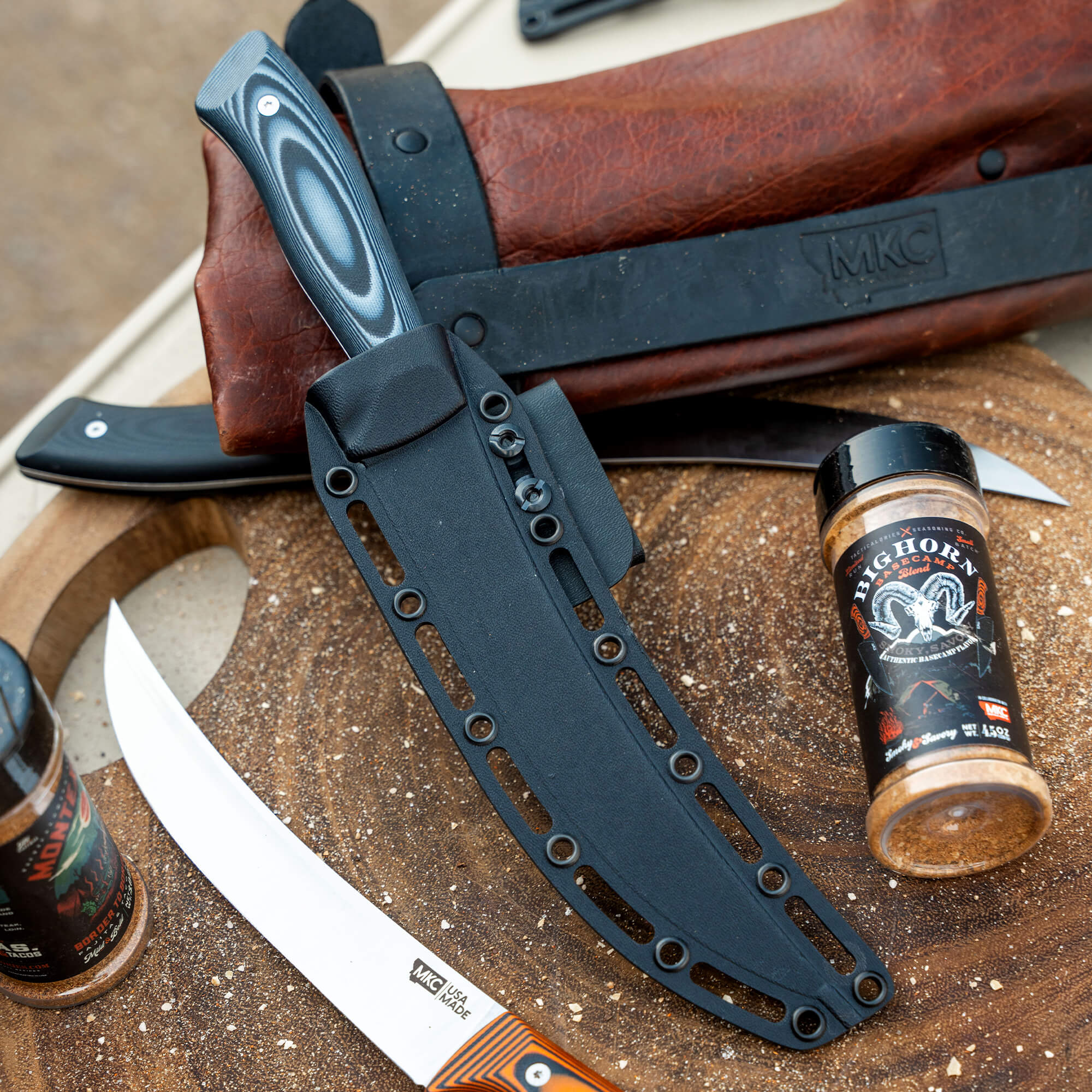 Product image featuring a collection of Bearded Butchers items including a sheathed black tactical knife, various unsheathed butcher knives with wooden handles, and a bottle of Bearded Butchers seasoning on a rustic wooden surface.