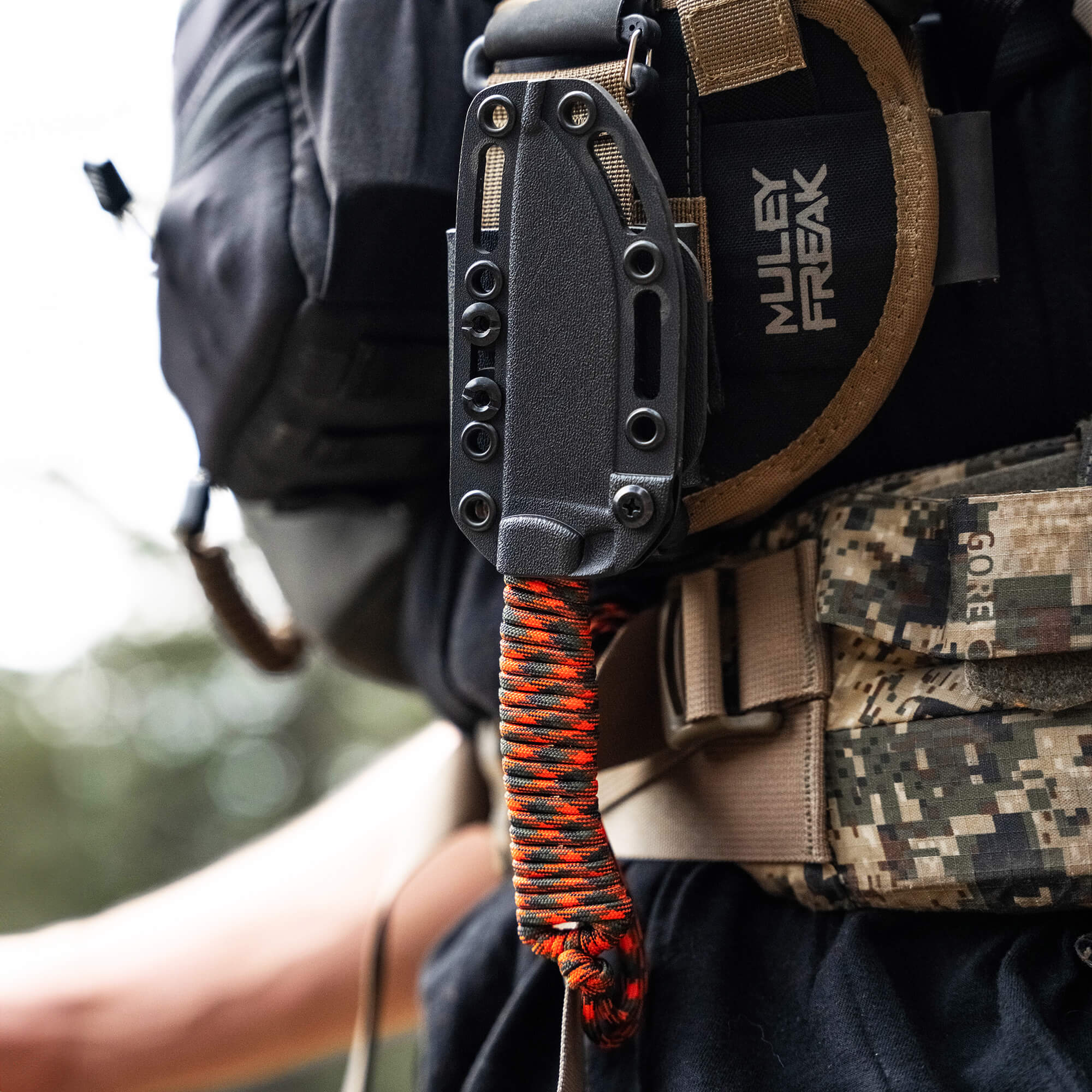 A tactical knife with black sheath and orange-and-black paracord attached to a camouflage backpack, indicating outdoor utility and readiness.