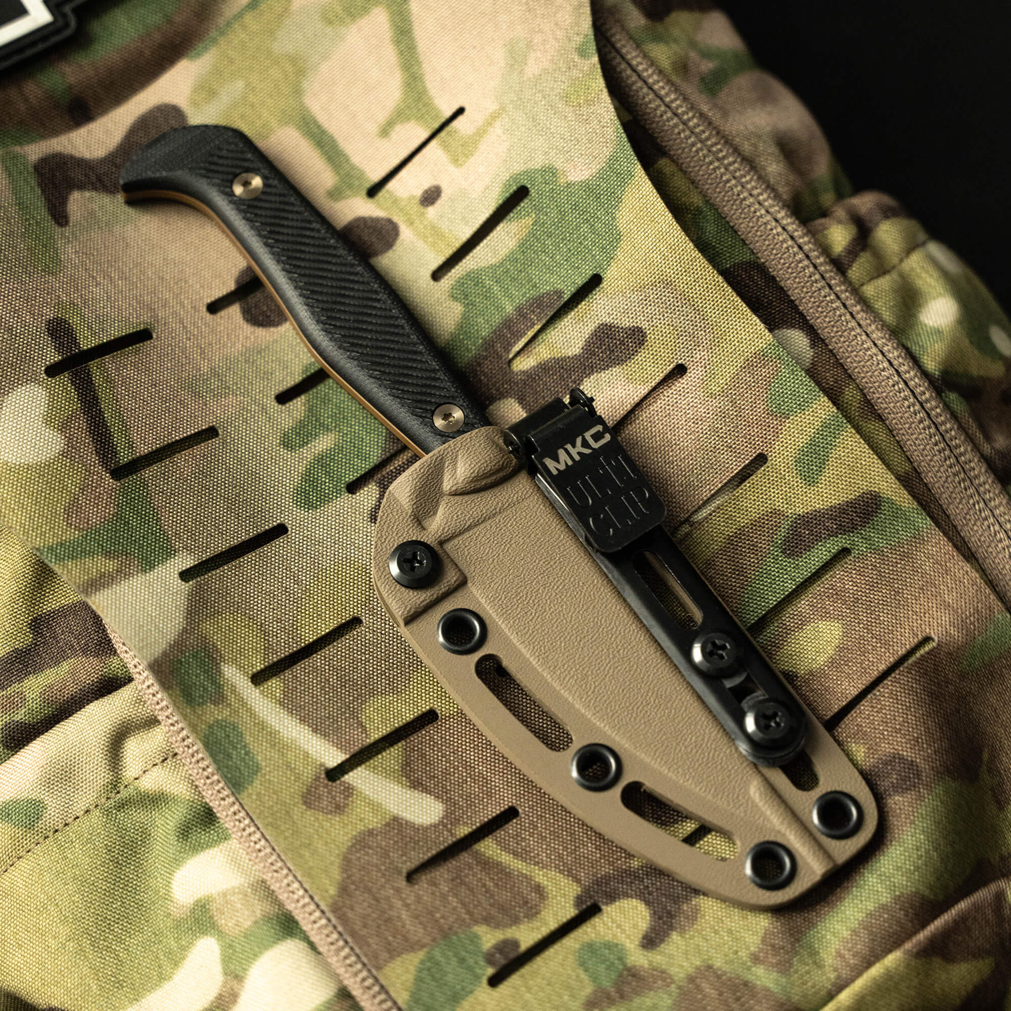 Alt text: "Close-up view of a BATTLE GOAT tactical knife in Coyote Black colorway, featuring a textured handle and black sheath, secured to a camouflaged tactical backpack with MOLLE webbing."