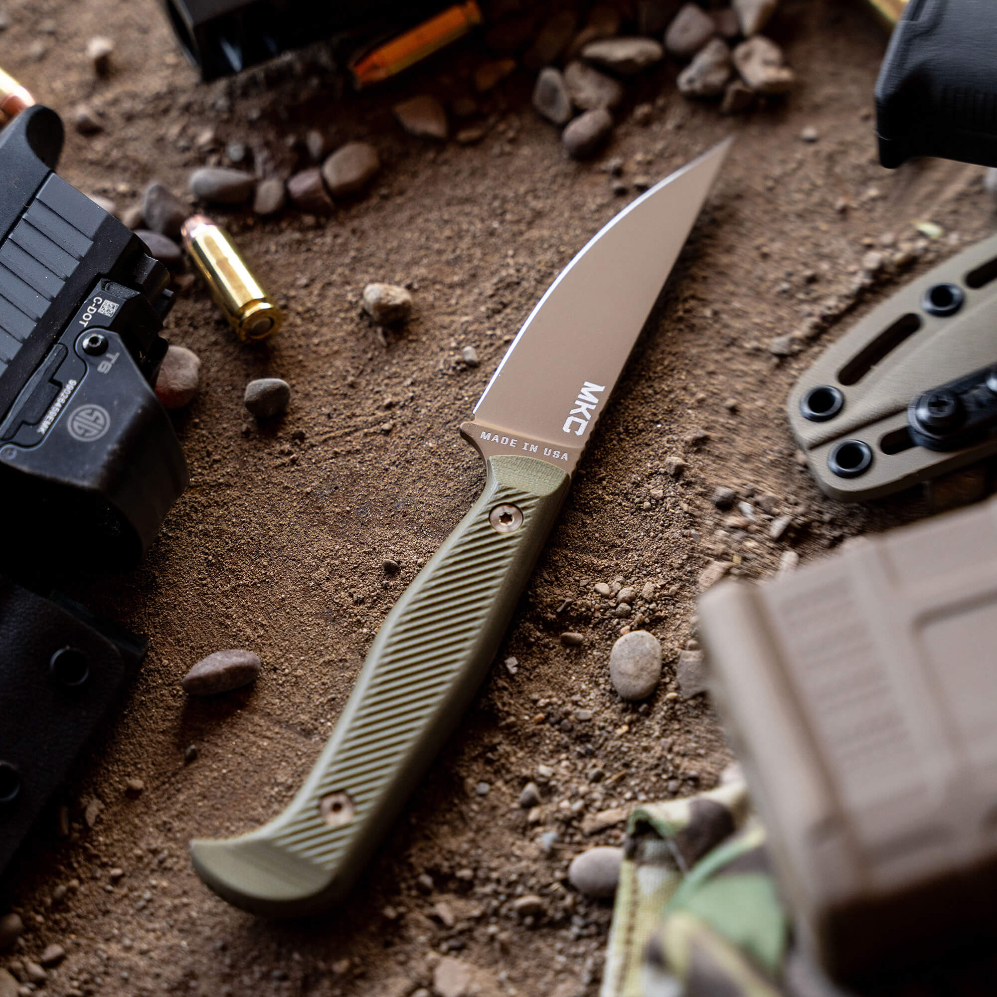 Alt text: "Close-up of a Battle Goat tactical knife with a coyote olive drab handle laid on a sandy surface surrounded by tactical gear including a handgun, bullets, and military equipment."