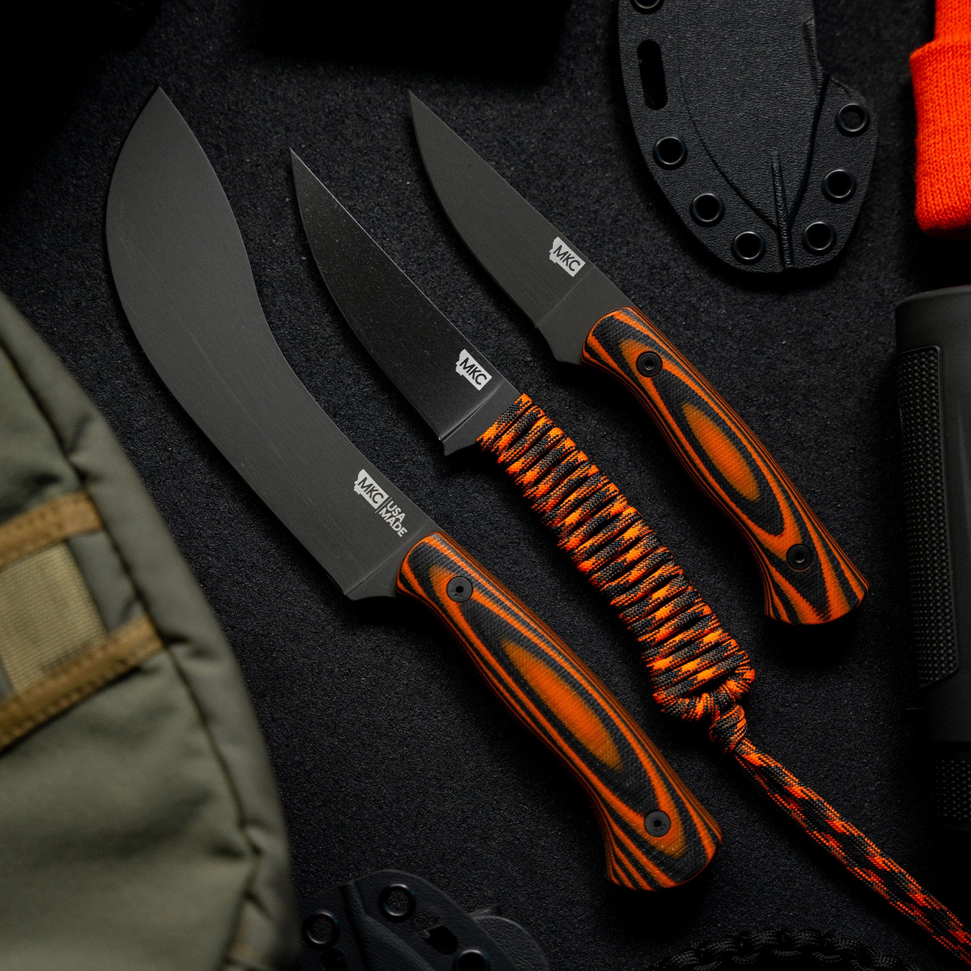 Alt text: "A set of three Beartooth professional skinning knives with orange and black handles, displayed on a dark textured background. The knives vary in size and include paracord-wrapped handles and black blades, accompanied by sheaths."