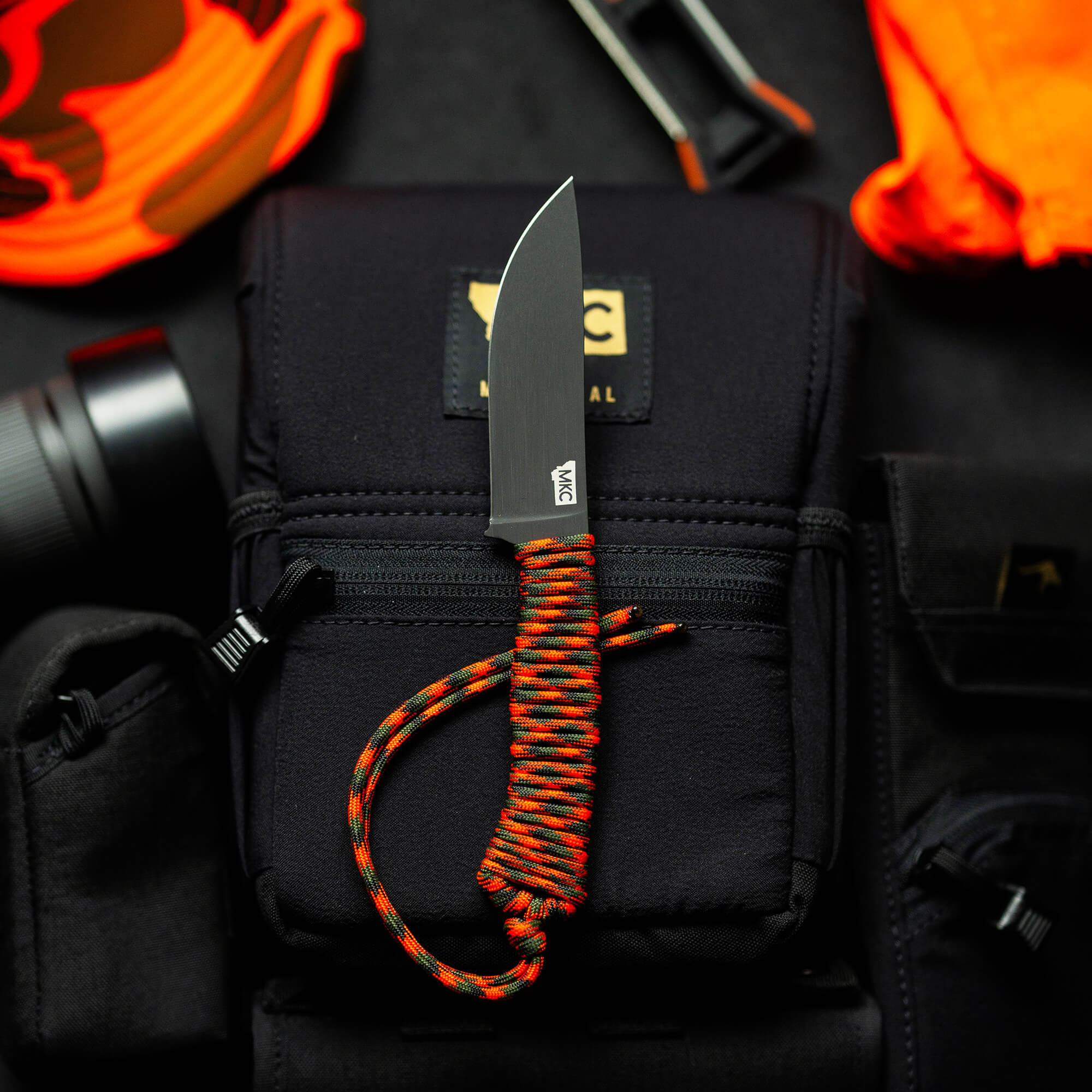 Alt text: "Sturdy tactical knife from Wild Sheep Foundation Stoned Goat 2.0, featuring a sharp, straight blade and vibrant orange and red paracord handle, displayed on a black tactical pouch with various outdoor gear in the soft-focus