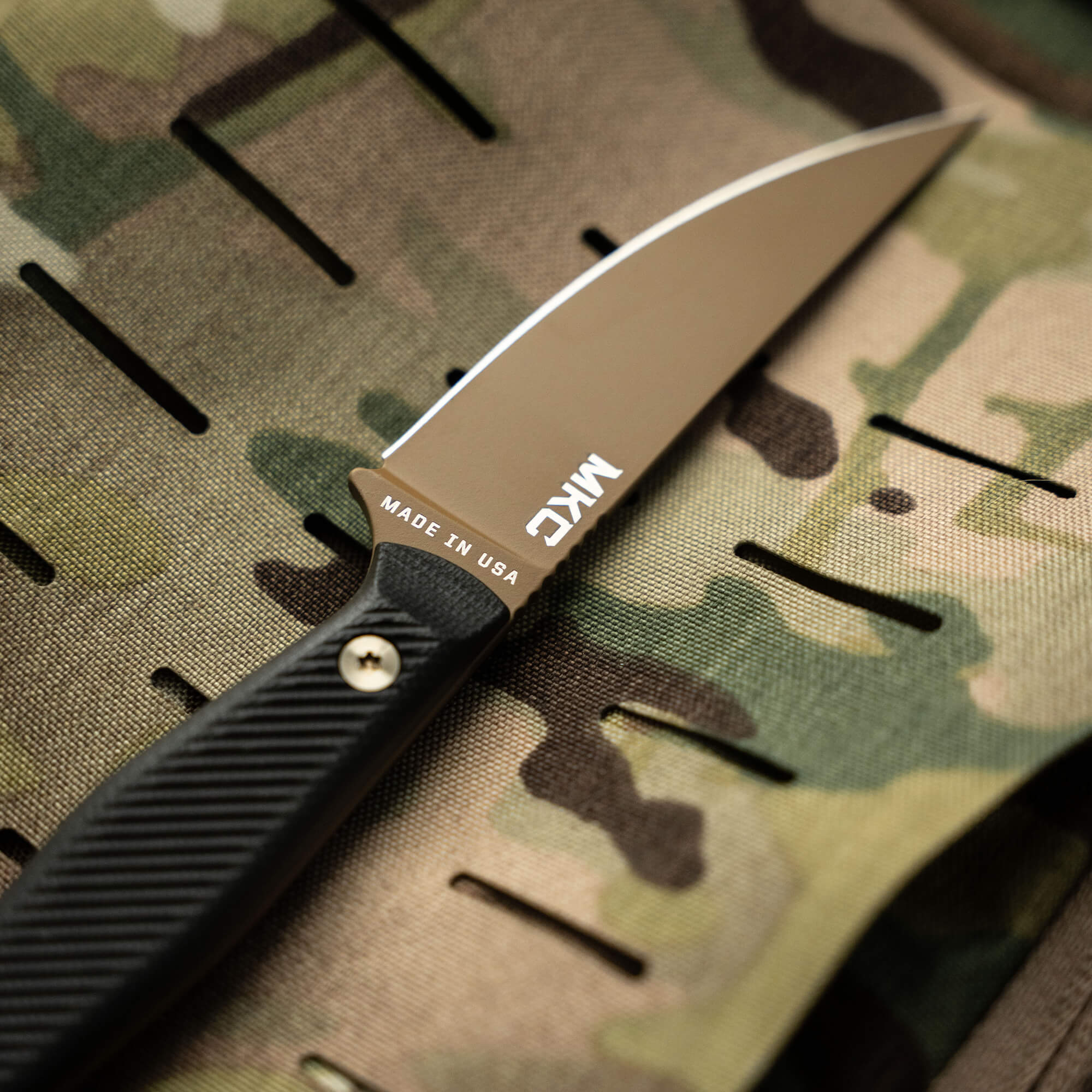 Alt text: "Close-up view of the BATTLE GOAT tactical knife with a COY/BLK color scheme, featuring a sleek black handle and silver blade marked 'Made in USA,' resting on a camouflage background."