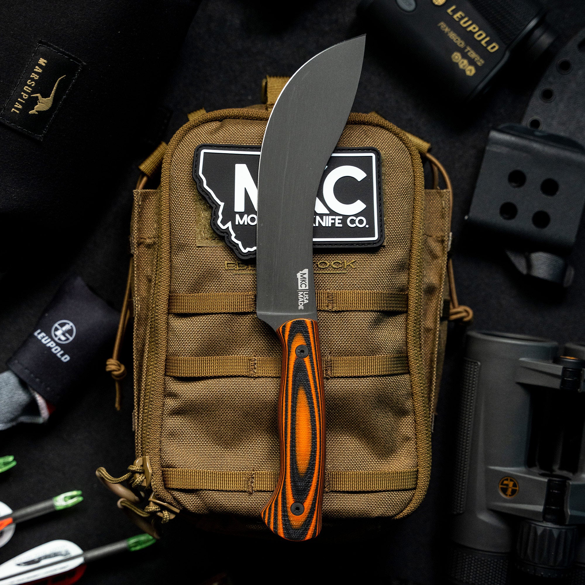 Alt text: "Beartooth Pro Skinning Blade with an orange and black handle prominently displayed on a tactical brown bag with patches, surrounded by outdoor gear including binoculars and a camera, highlighting the knife's rugged use."