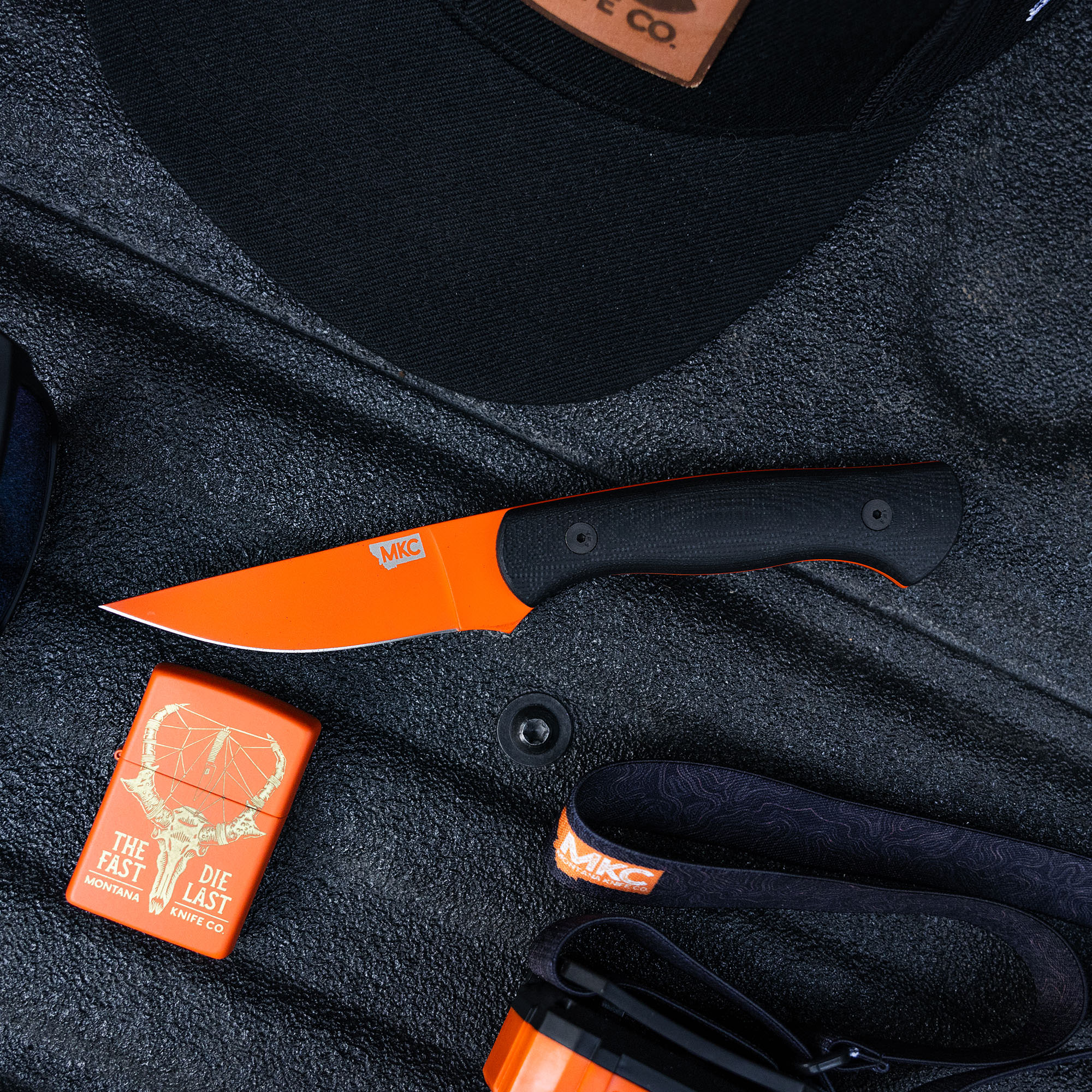 Blaze orange Blackfoot 2.0 knife with black handle and brand logo on dark fabric background accompanied by a branded lighter and cap.