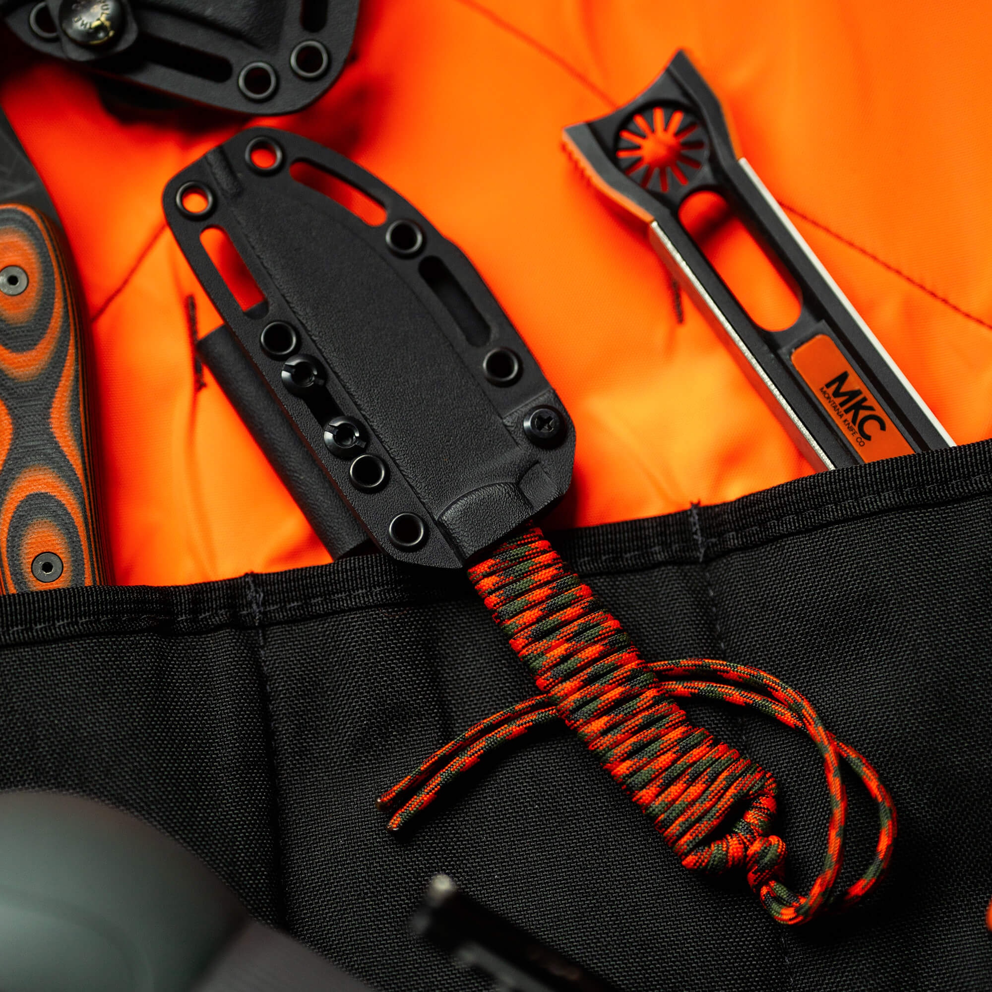 Alt text: Close-up view of the MKC Stoned Goat 2.0 knife from the Wild Sheep Foundation secret drop, featuring a black sheath and orange and black handle, contrasted against a bright orange jacket with a paracord accessory