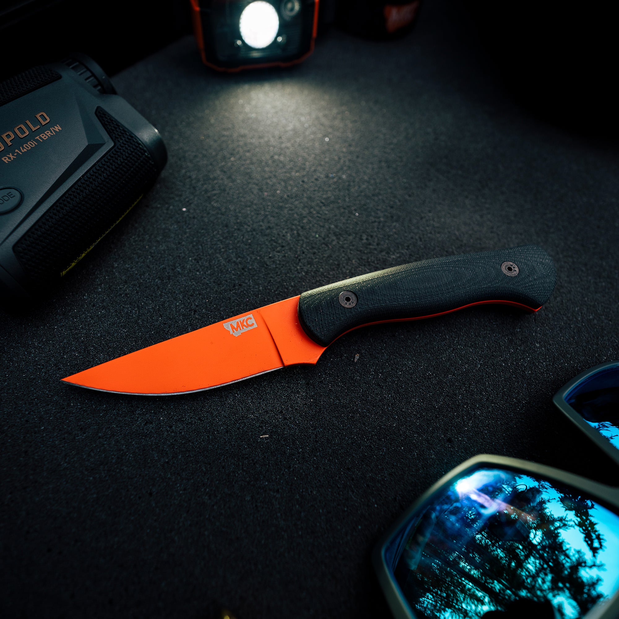 Alt text: "BLACKFOOT 2.0 fixed blade knife with a blaze orange Cerakote™ blade and black handle, displayed on a dark surface next to outdoor gear."