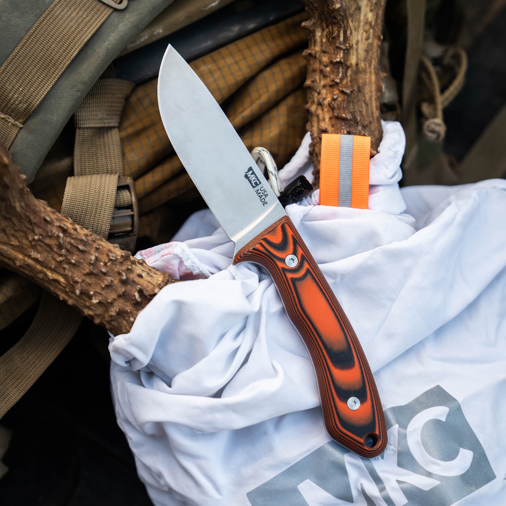 MKC Mule Deer Knife in orange and black, resting on a white bag among hunting gear and deer antlers.