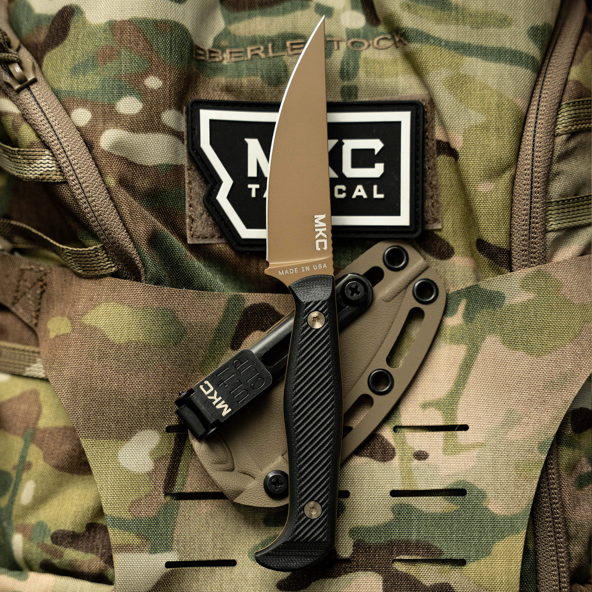 Alt text: "Battle Goat tactical knife in coyote black with a fixed blade displayed on a camouflaged backpack, featuring MKC Tactical logo and 'Made in USA' tag."