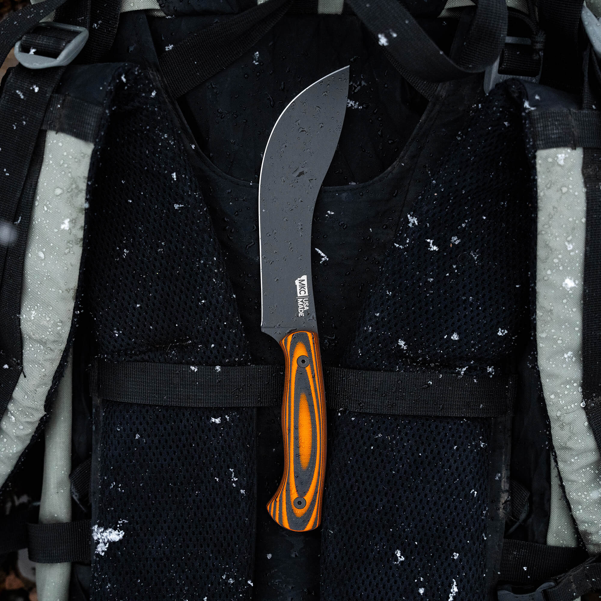 Alt text: "Beartooth Pro Skinning Blade with a curved black blade and vibrant orange and black handle, secured on a black tactical vest with visible snowflakes."
