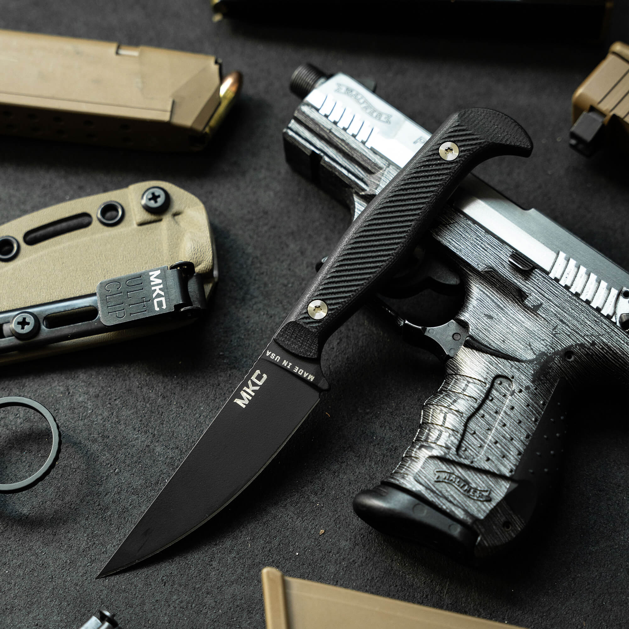 Alt text: Close-up view of a Mike Battle Goat folding knife in black, with a distinctive textured handle and sleek blade, displayed alongside tactical gear including a flashlight and folding tool on a dark, textured surface.