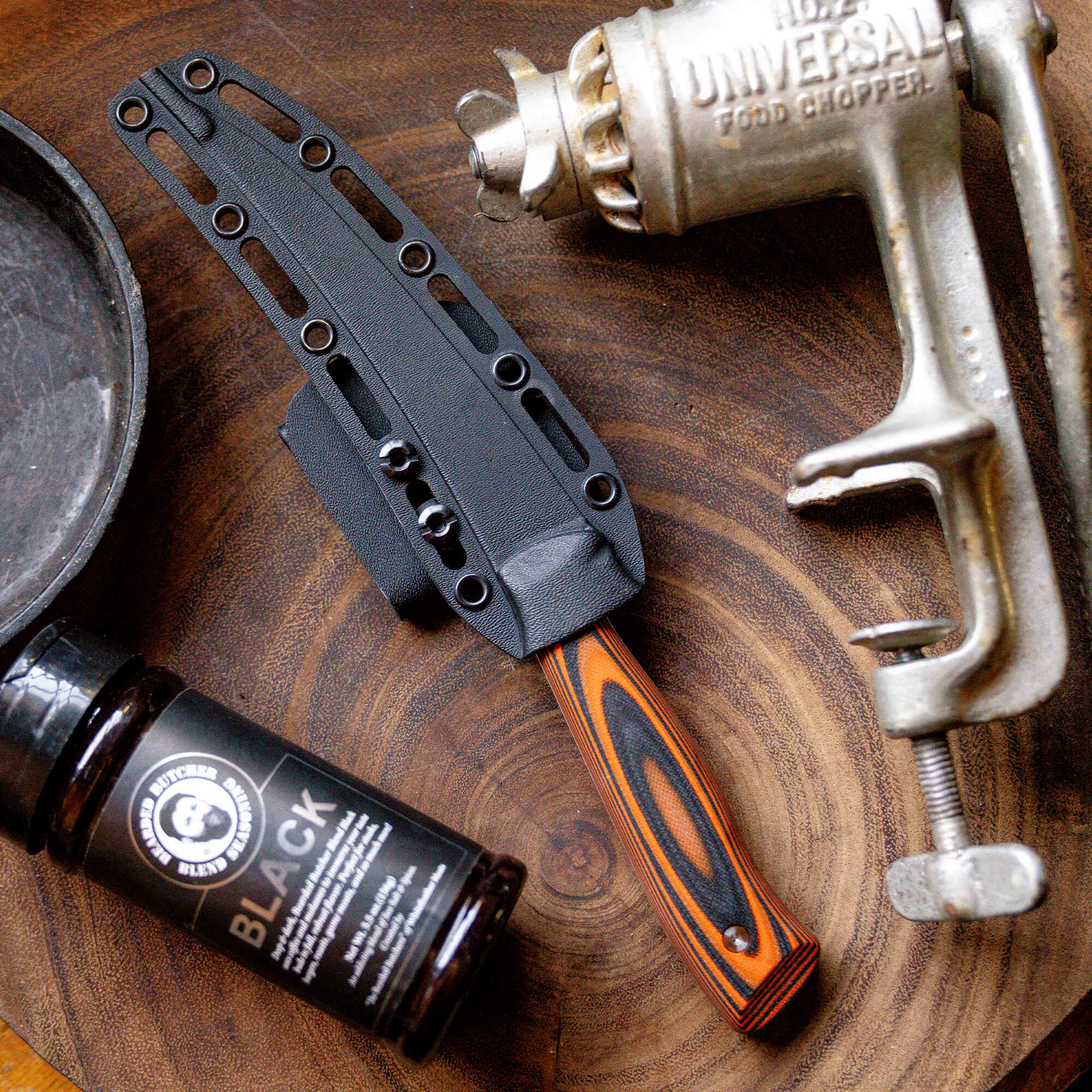 Alt text: "Bearded Butchers boning butcher knife with black sheath and distinctive orange and black handle, displayed next to a bottle of Bearded Butcher blend seasoning and an antique food chopper, all arranged on a wooden surface."