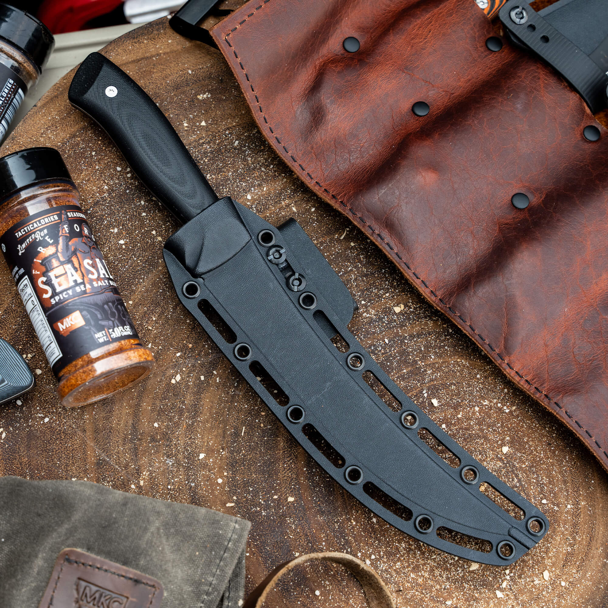 Alt text: "A robust breaking butcher knife from Bearded Butchers with black handle and sleek sheath, displayed alongside a bottle of seasoning sauce and a rustic leather apron on a textured surface, indicating a sold-out secret product drop."