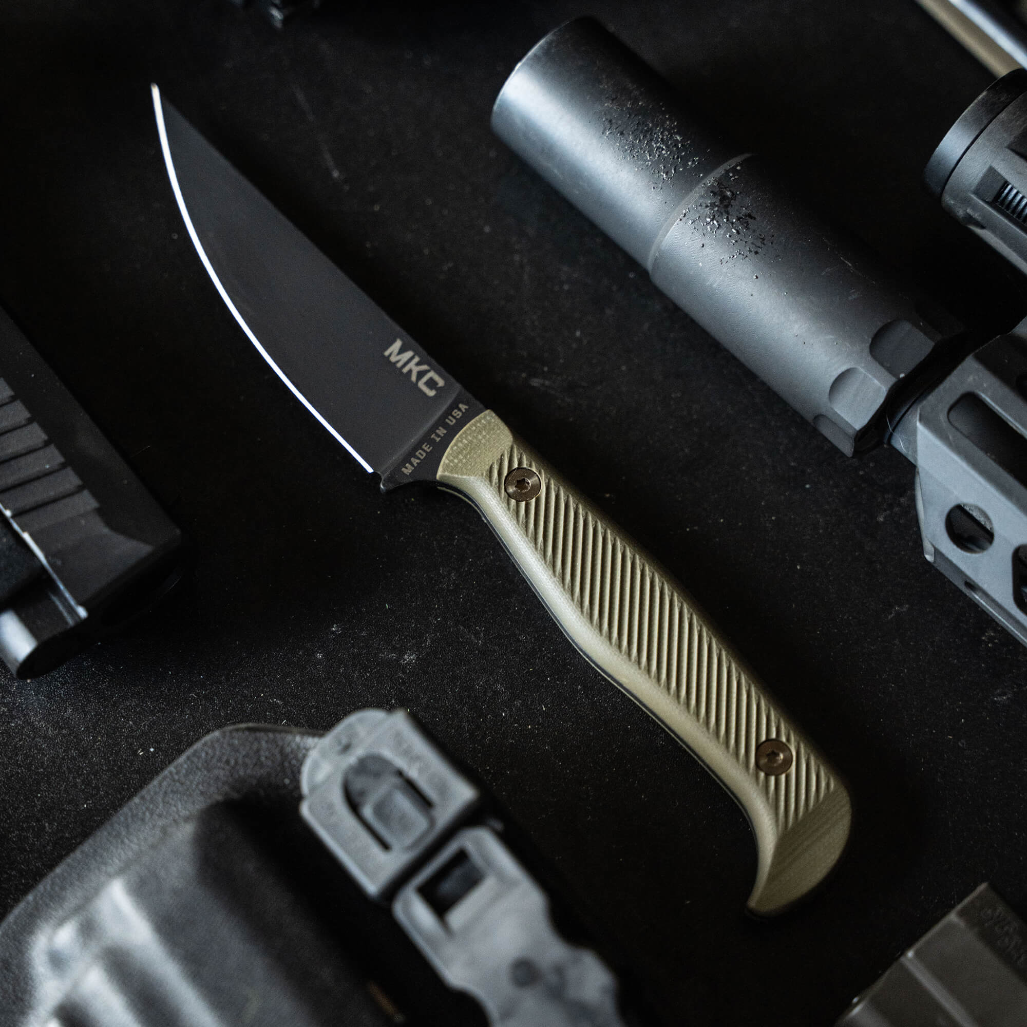 Alt text: Black MKC BATTLE GOAT knife with olive drab handle displayed among tactical gear, featuring a sharp blade and textured grip.