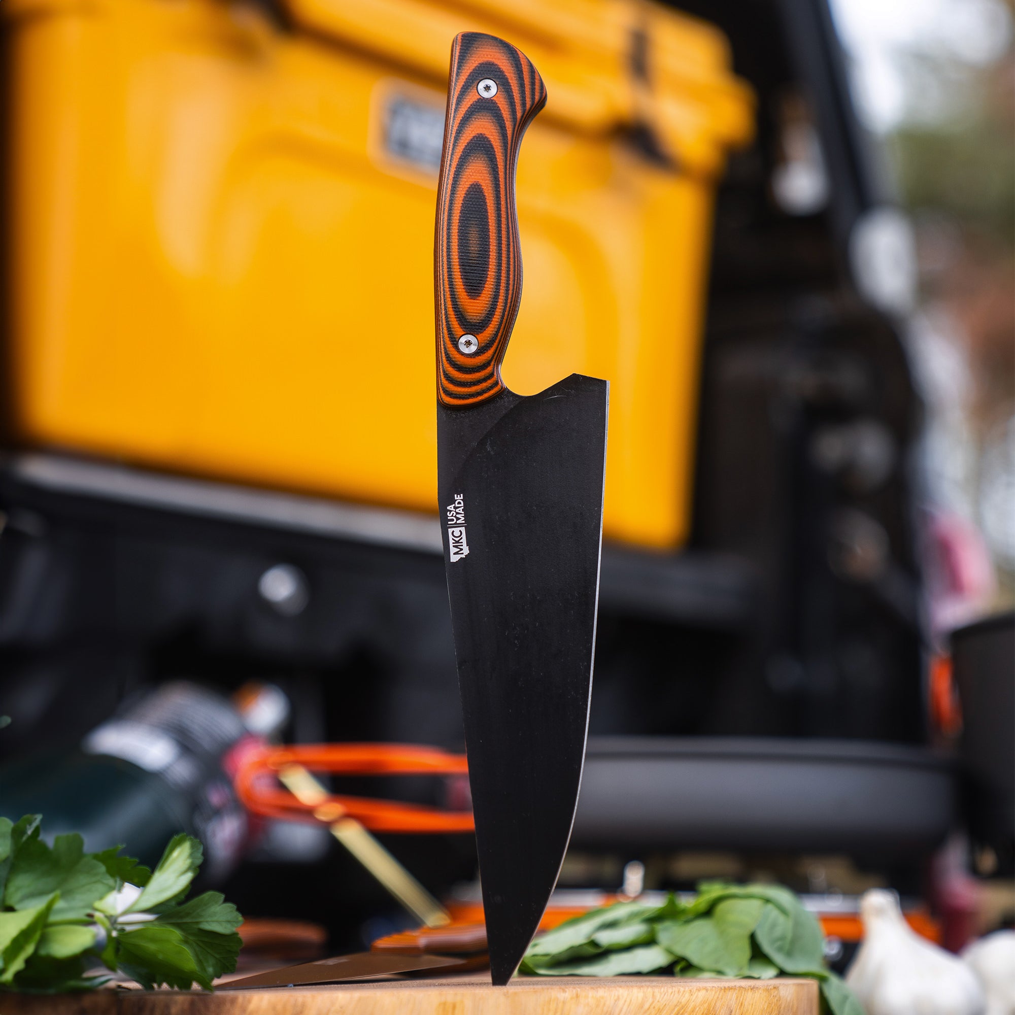 Alt text: "BIGHORN CHEF cleaver knife with orange and black handle, prominently displayed with its blade lodged vertically in a wooden cutting board, set against a backdrop of a yellow vehicle, outdoors."