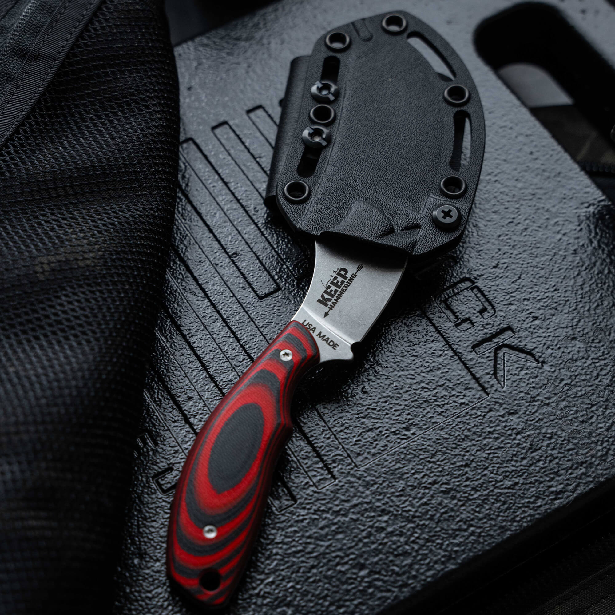 Close-up of a Packout Skinner knife with a red and black handle partially sheathed in a black Kydex holster, resting on a textured black surface with embossed logos.