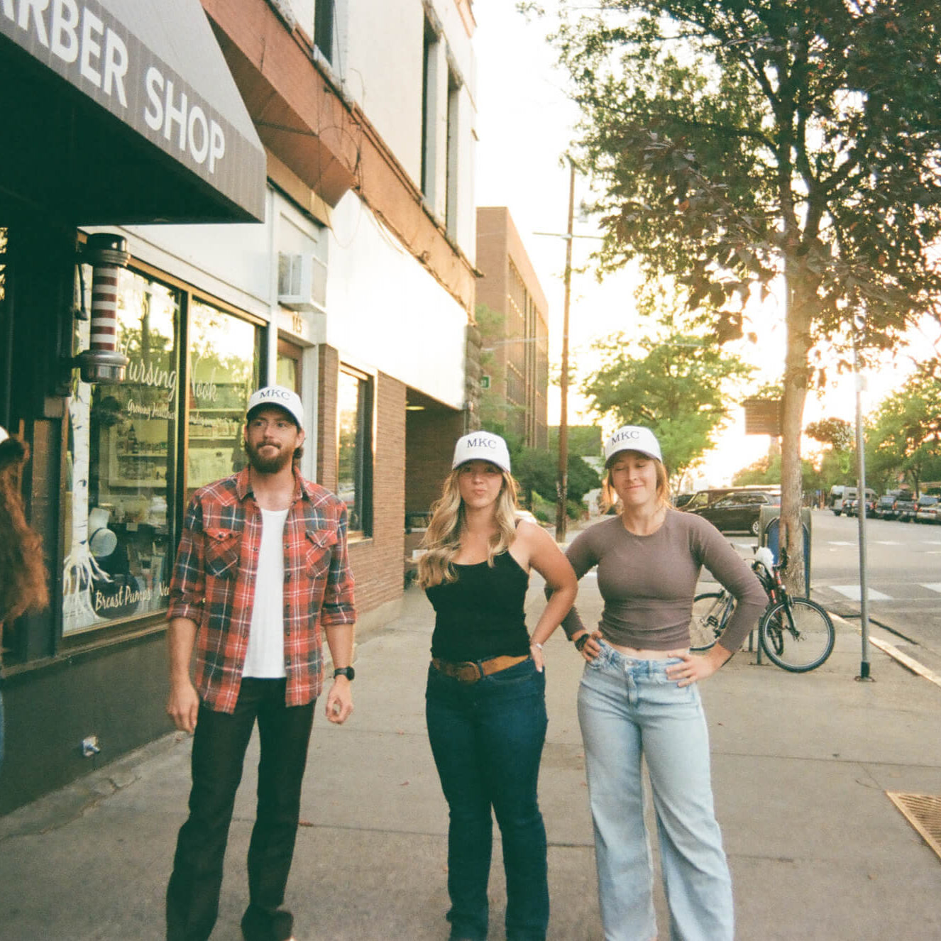 Four people standing on a sidewalk in front of a barber shop with trees and cars in the background.