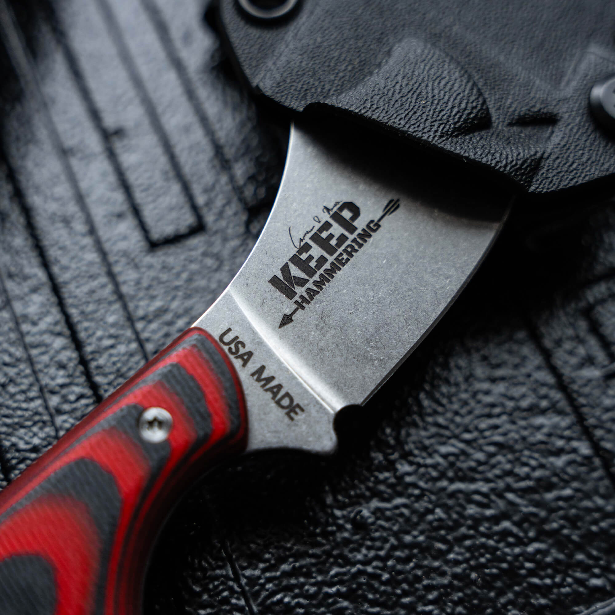 Close-up image of the "Keep Hammering Packout Skinner" knife, showcasing its engraved blade with the "Keep Hammering" logo and red and black handle, lying on a textured black surface.