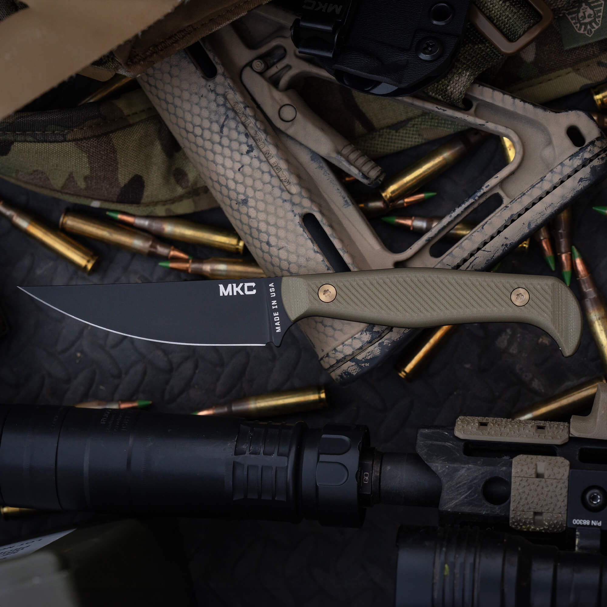 Alt text: "Close-up image of the MKC Battle Goat BLK/OD tactical knife with a serrated black blade and olive drab green handle, displayed among military gear and ammunition."