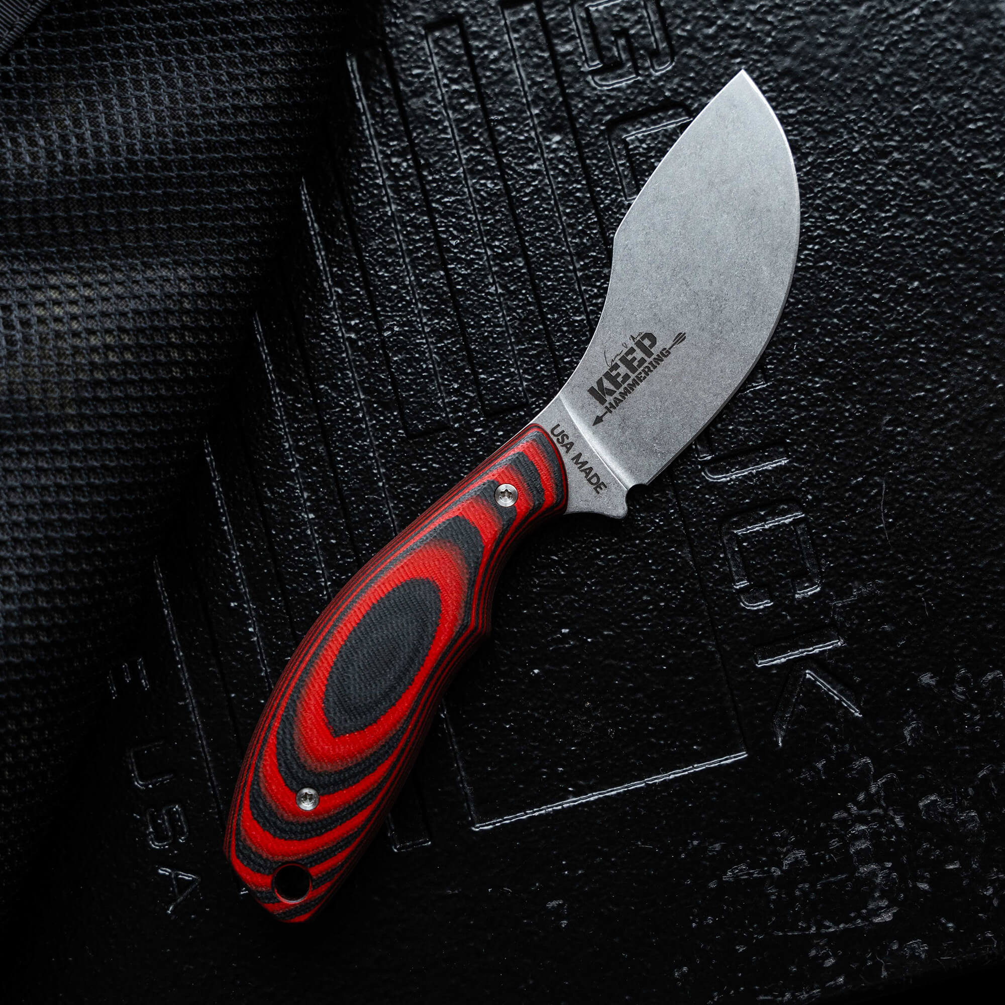 Alt text: Close-up of a Keep Hammering Packout Skinner knife with a red and black handle, showcasing the blade etched with "KEEP" and "US-MADE" on a textured black background.