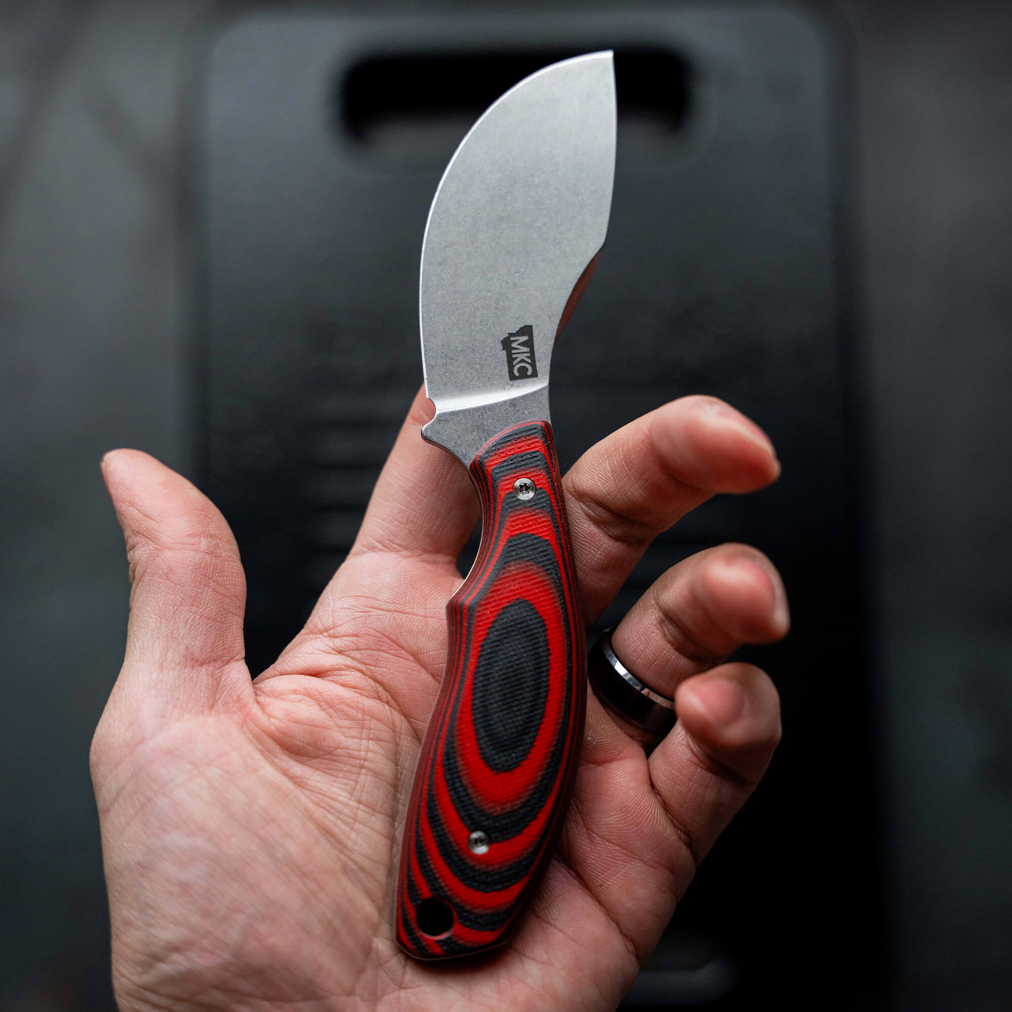 Alt text: "Close-up of a hand holding a limited edition 'Keep Hammering Packout Skinner' knife with a red and black handle and a matte finished blade, against a soft black background."