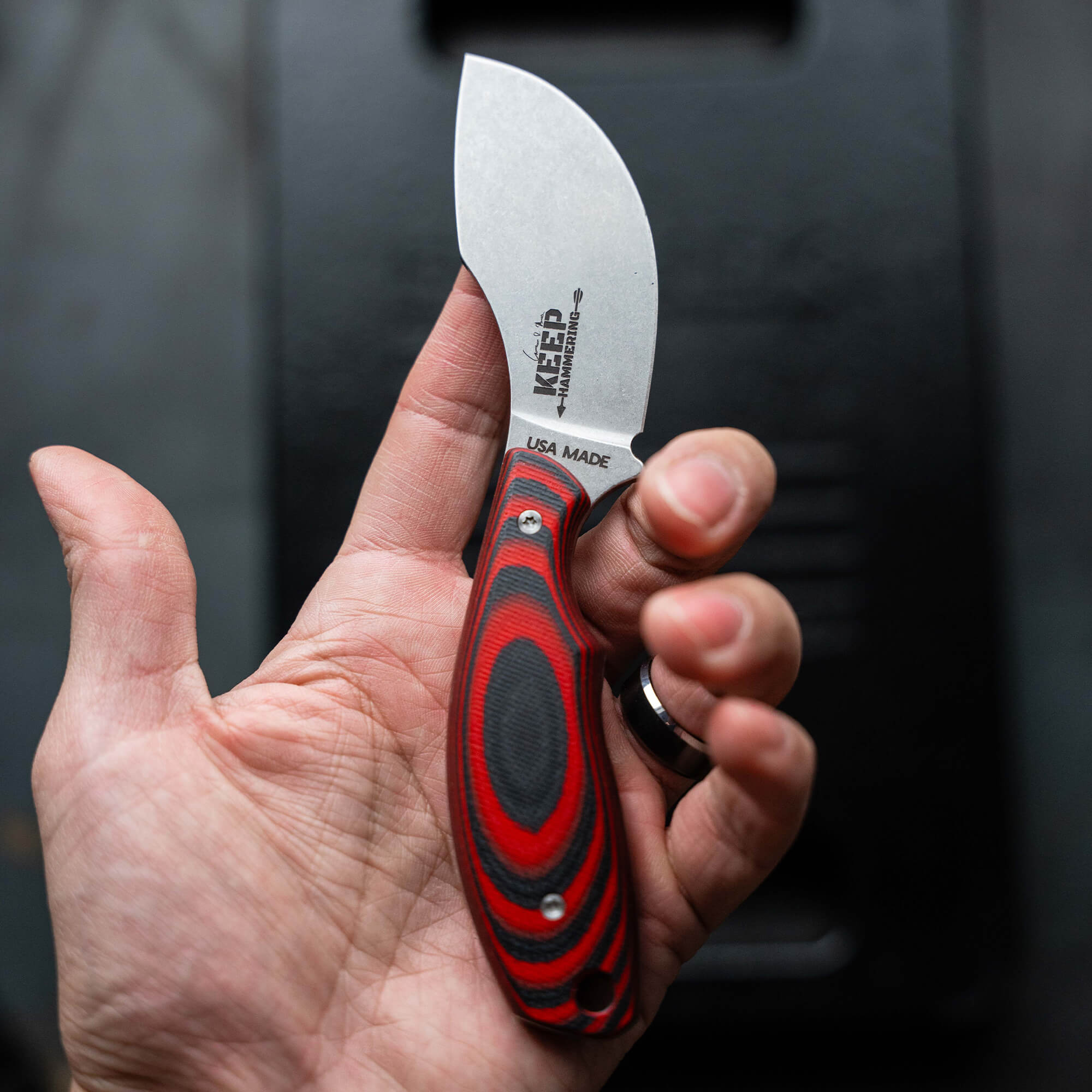 Alt text: Close-up of a hand holding a "Keep Hammering Packout Skinner" knife, featuring a red and black grip and a broad, curved stainless steel blade with the logo and "USA MADE" text. Background includes a black textured case
