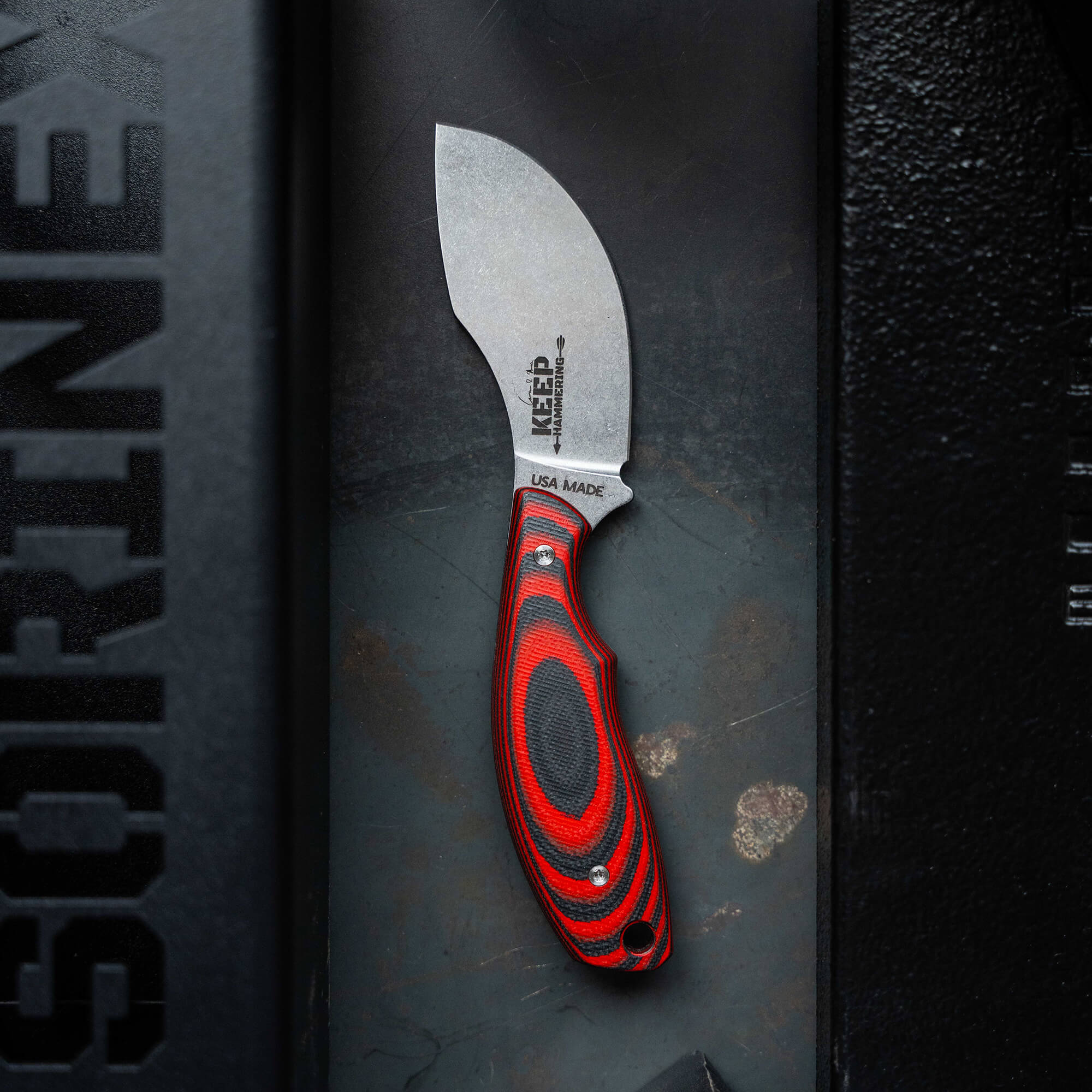 USA-made skinner knife with a vibrant red and black ergonomic handle, displayed against a textured dark background, highlighting the "Keep Hammering" and "Packout" brand details on the blade.