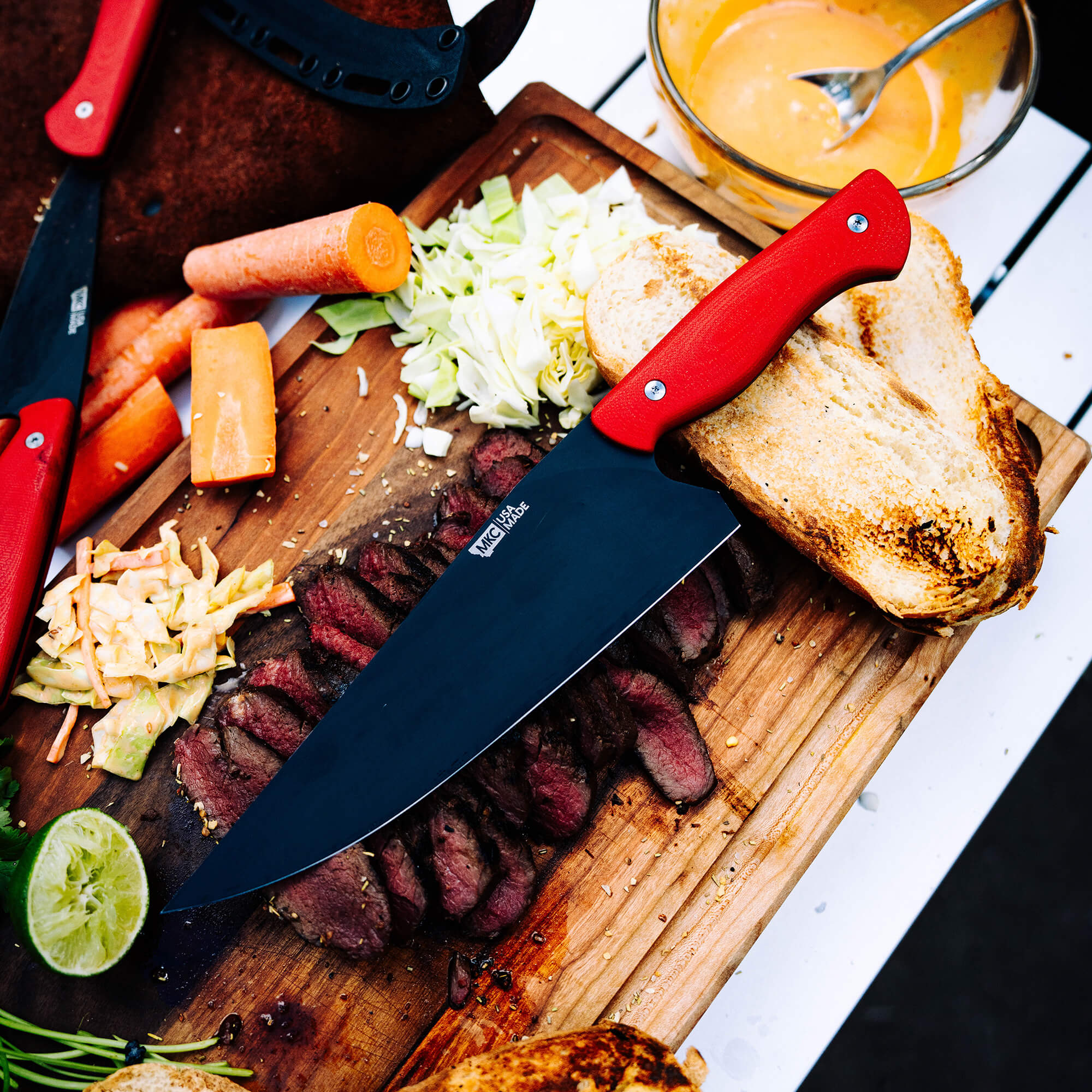 Alt text: "Top view of a BIGHORN CHEF kitchen knife with a vibrant red handle, prominently resting on a wood cutting board alongside ingredients including sliced carrots, cabbage, grilled bread, and cooked steak slices."