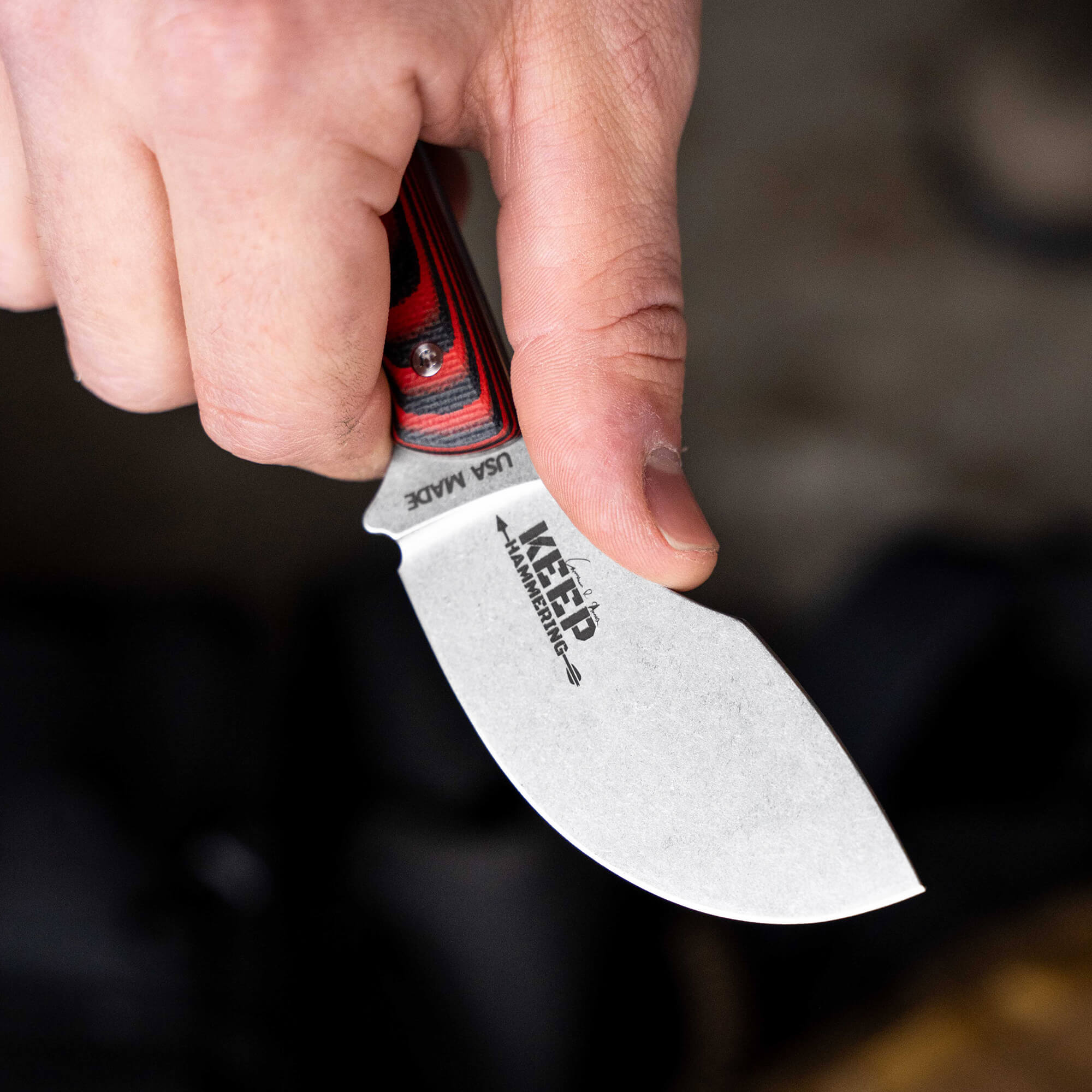 Close-up of a hand holding a "Keep Hammering - Packout Skinner" knife with a red and black layered handle, showcasing the blade's unique shape and engraved logo.