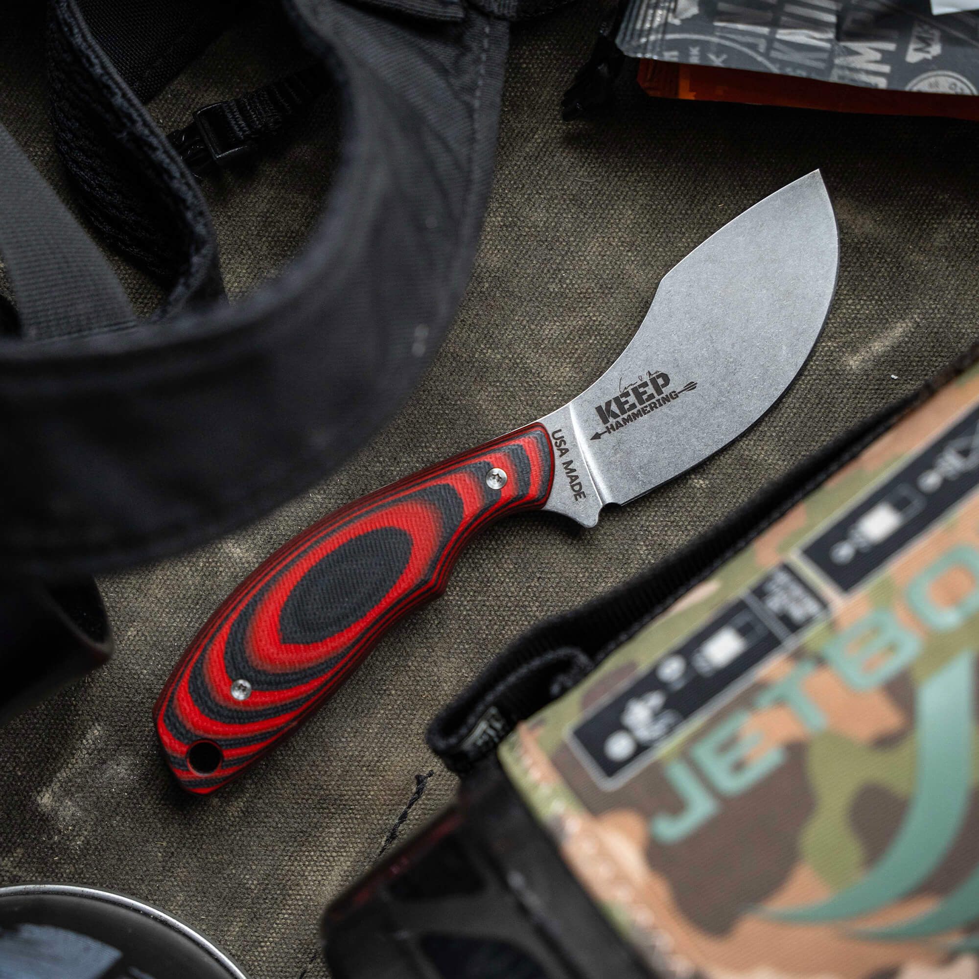Close-up image of a Keep Hammering Packout Skinner knife with red and black handle, laying on a camouflaged surface next to outdoor gear.
