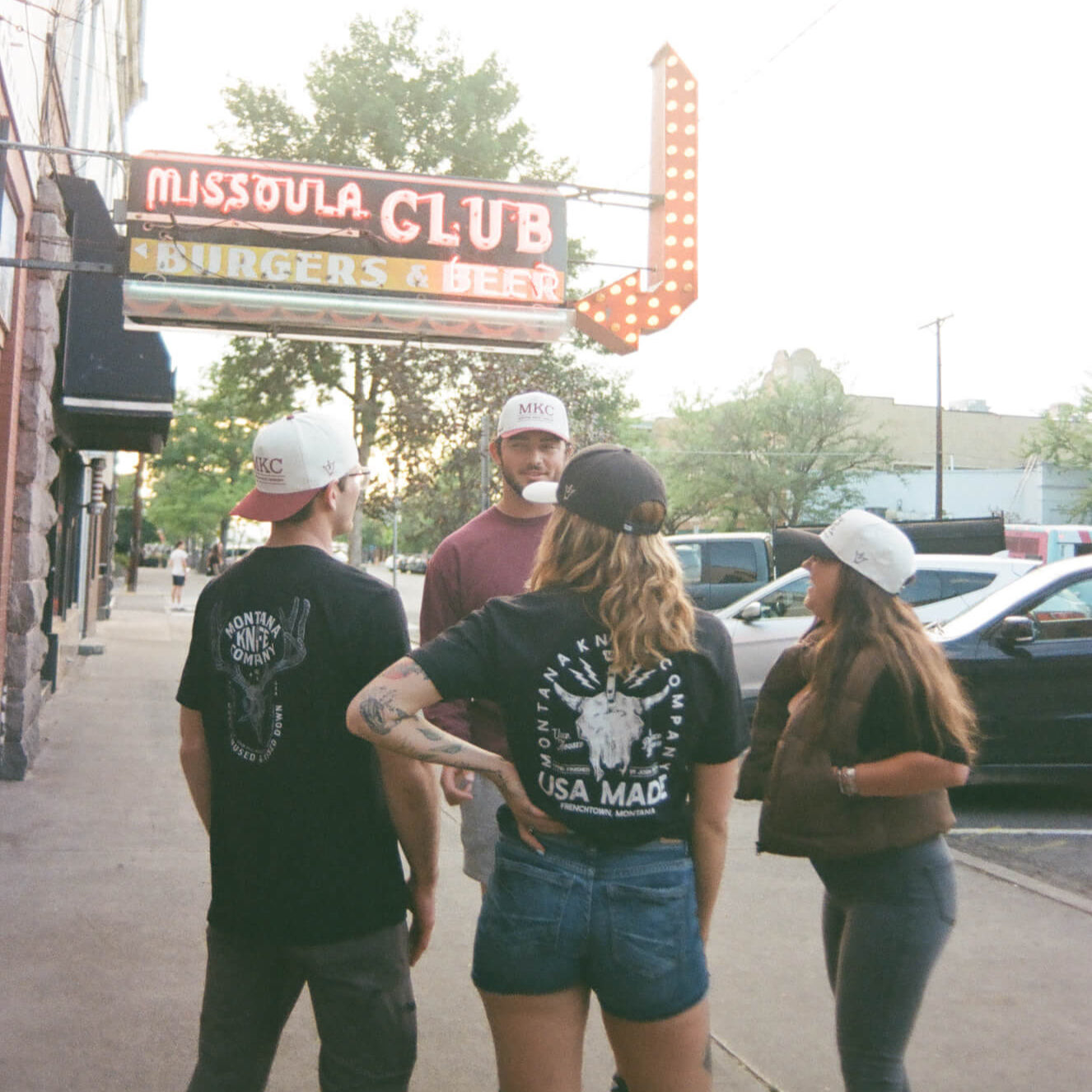 Four people standing on a sidewalk in front of a building with a sign that reads 'Missoula Club'.