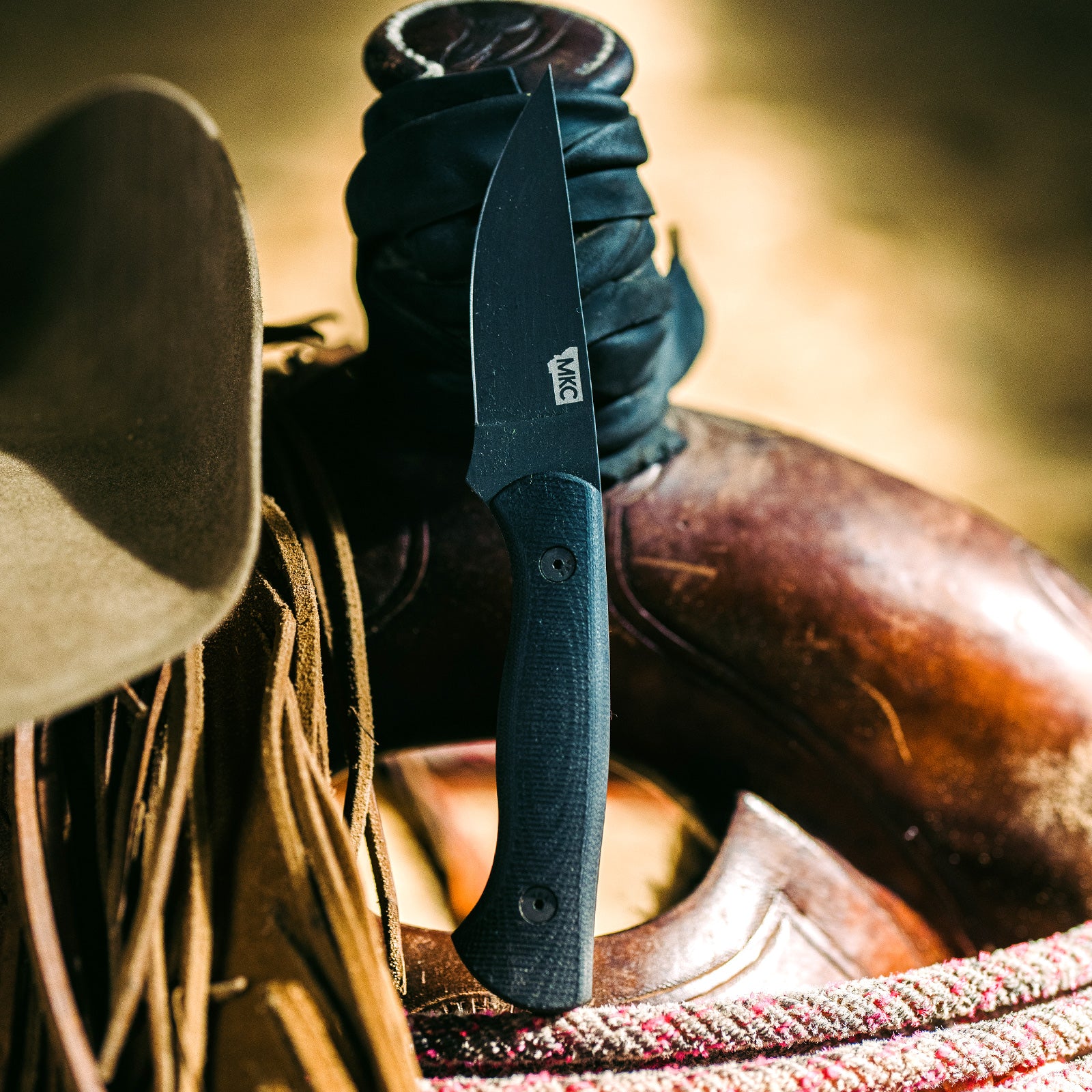 Alt text: "Close-up of the BLACKFOOT 2.0 tactical knife in black, featuring a textured handle and a sharp, angular blade, displayed against a rustic background with a leather saddle and cowboy hat."