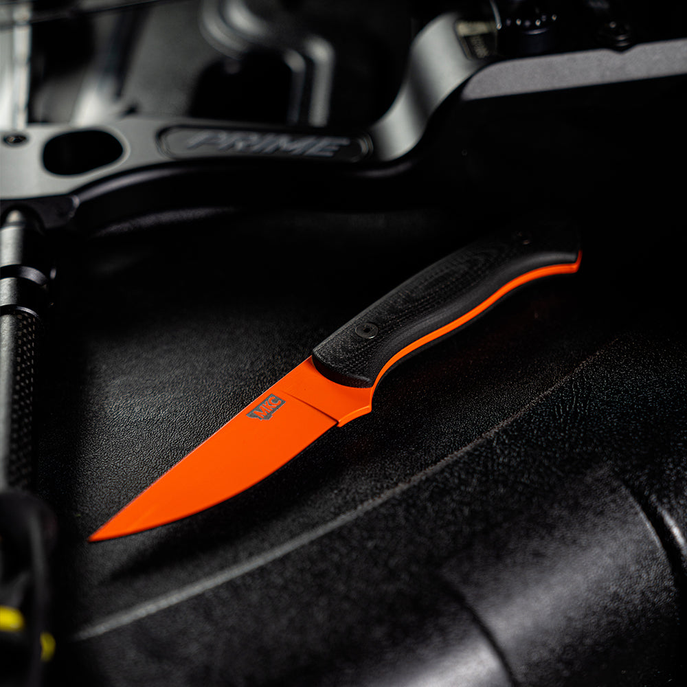 Alt text: "Close-up of a Blackfoot 2.0 fixed blade knife with a blaze orange Cerakote finish and black handle, resting on a textured black surface with what appears to be bike or mechanical parts in the background."