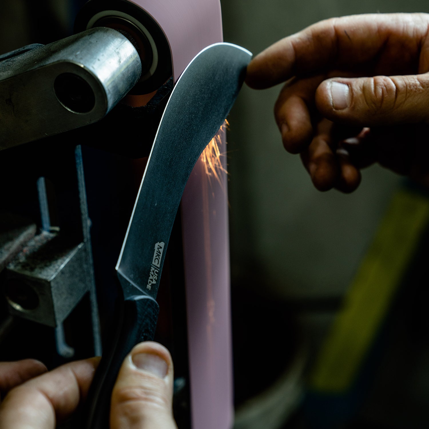 Alt text: Close-up of a person sharpening a Beartooth Pro Skinning Blade using a grinding wheel, with sparks flying off the blade in a workshop setting. The blade is prominently displayed with the "Beartooth" logo visible on its
