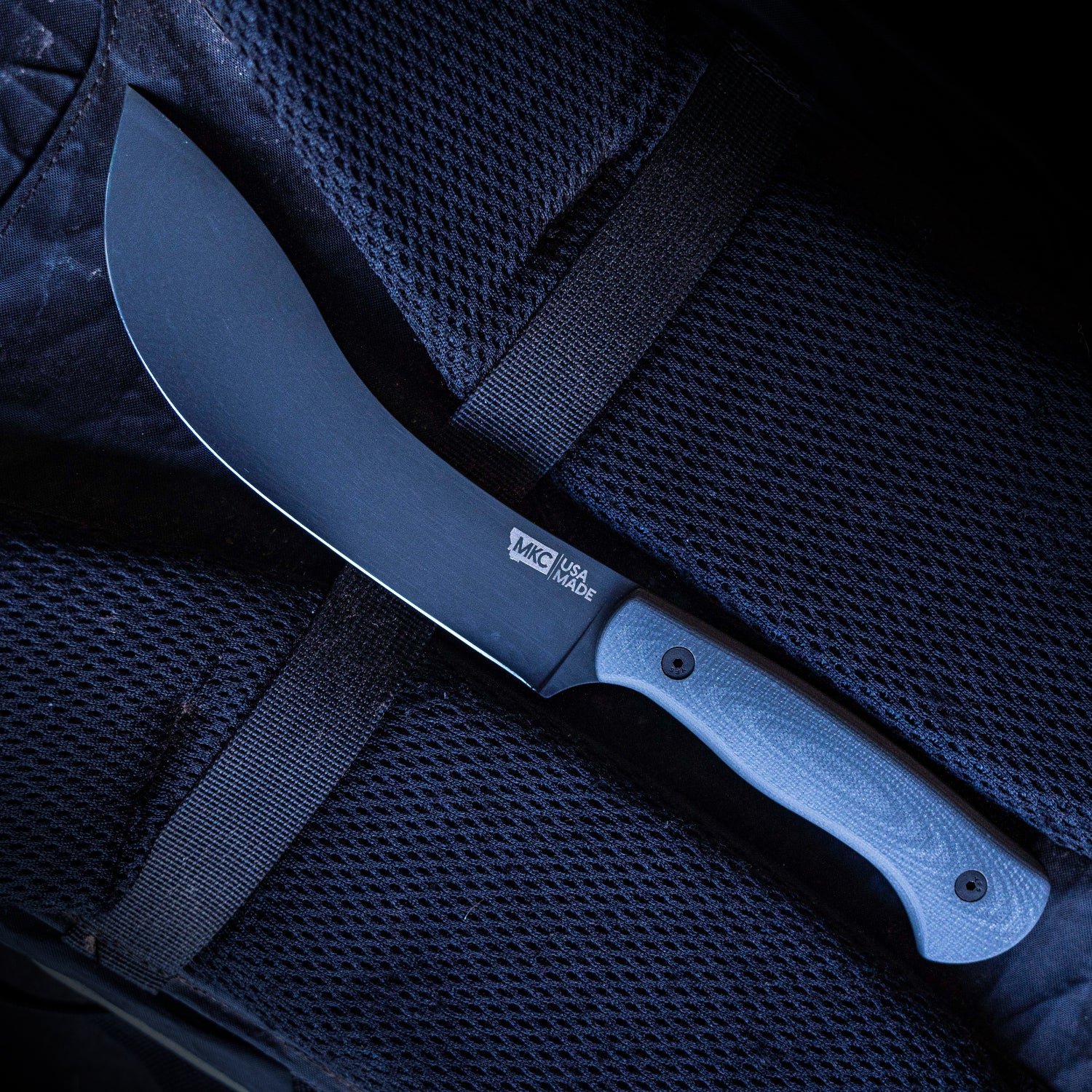 Alt text: "Close-up of a Beartooth Pro Skinning Blade with a grey handle, resting on a dark blue textured surface, showcasing the sharp curved blade and the etched brand logo MKC USA Made."