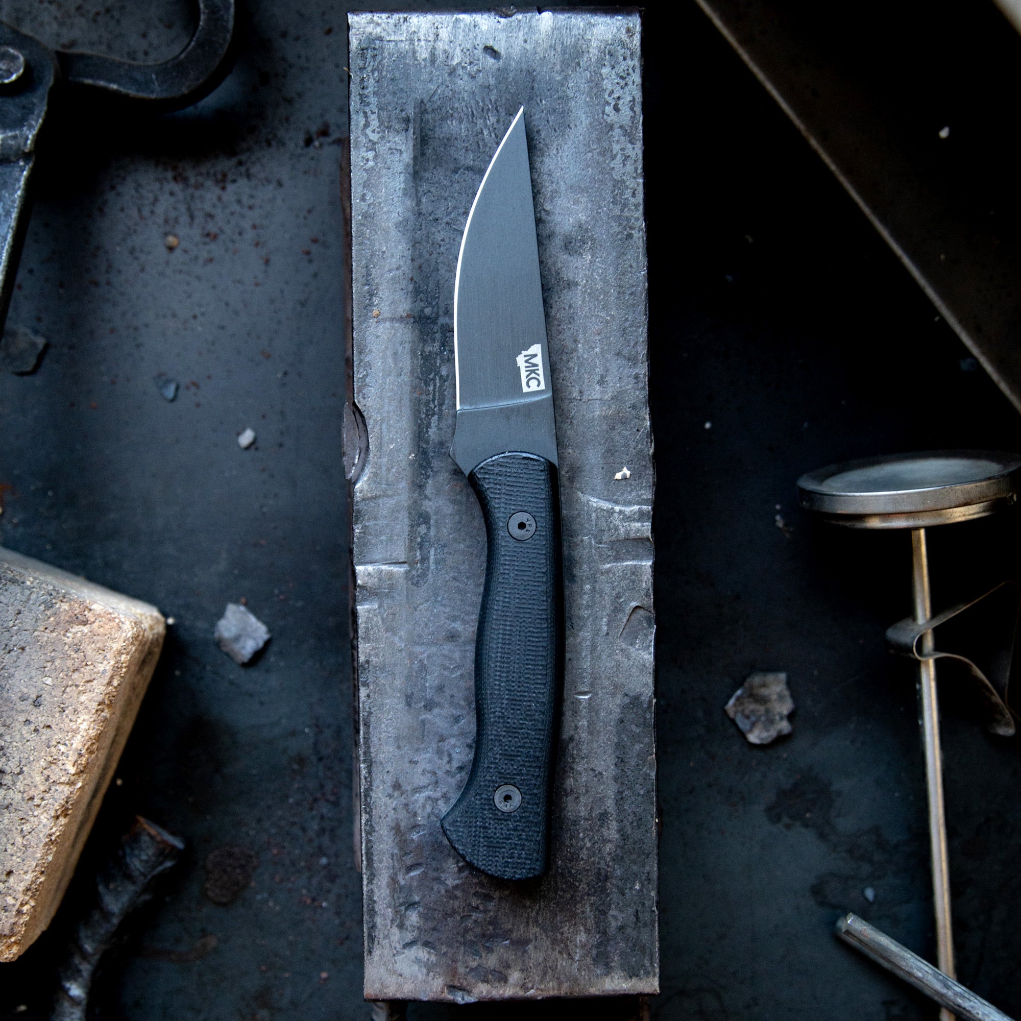 Alt text: "BLACKFOOT 2.0 tactical knife with a black blade and handle, displayed on a weathered metal surface in an industrial setting."