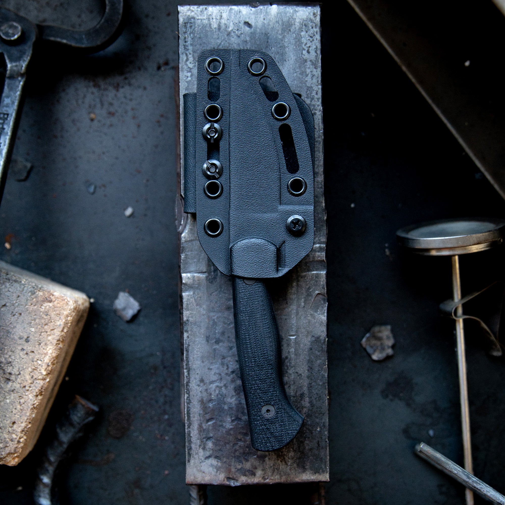 Alt text: "BLACKFOOT 2.0 tactical knife in black color with a textured handle and a custom-fit Kydex sheath, displayed on a gritty metal backdrop, emphasizing its robust and durable design."